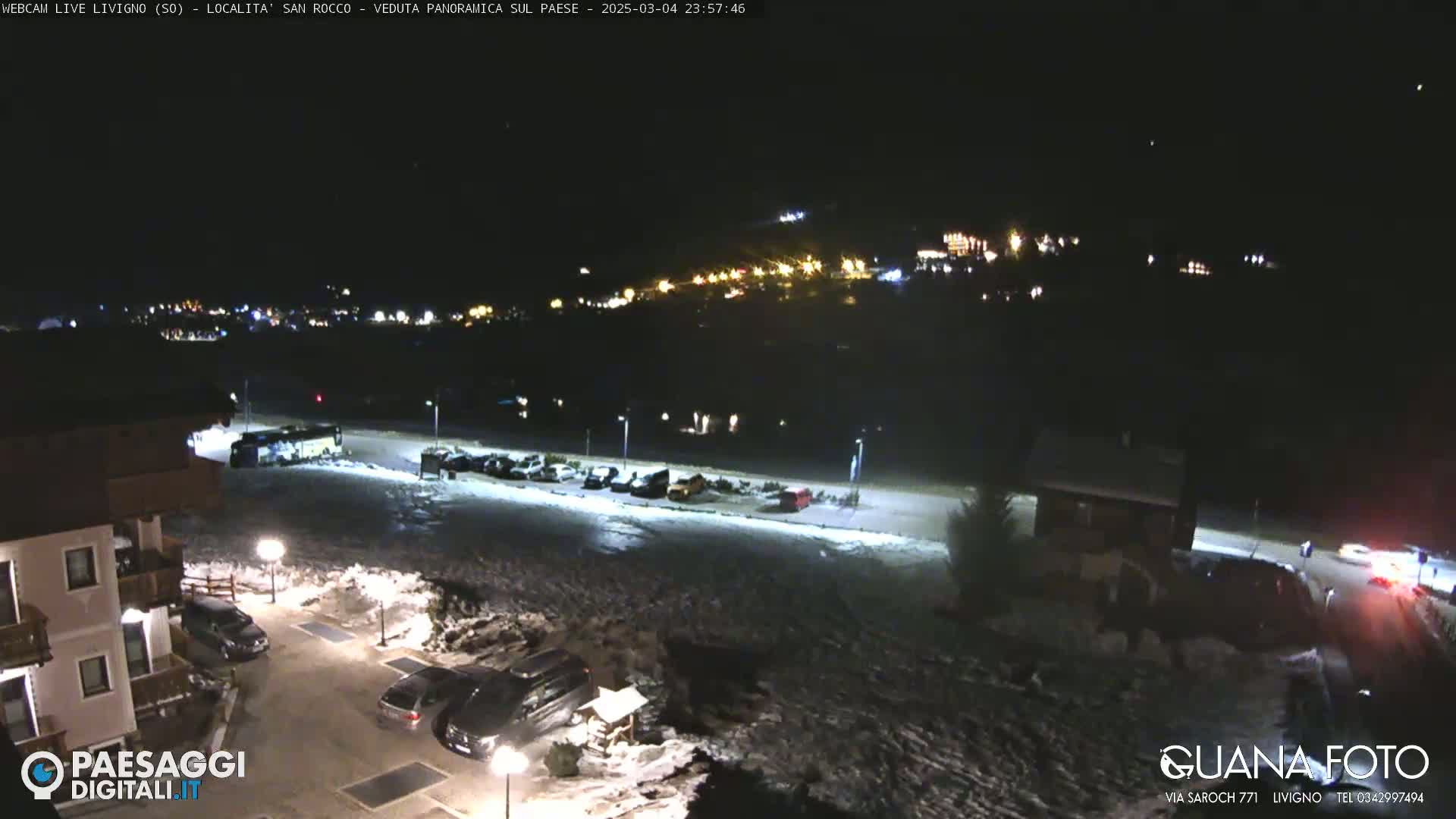 A nighttime, snow-covered parking lot and surrounding buildings are illuminated by streetlights, with a town's lights visible in the distance.