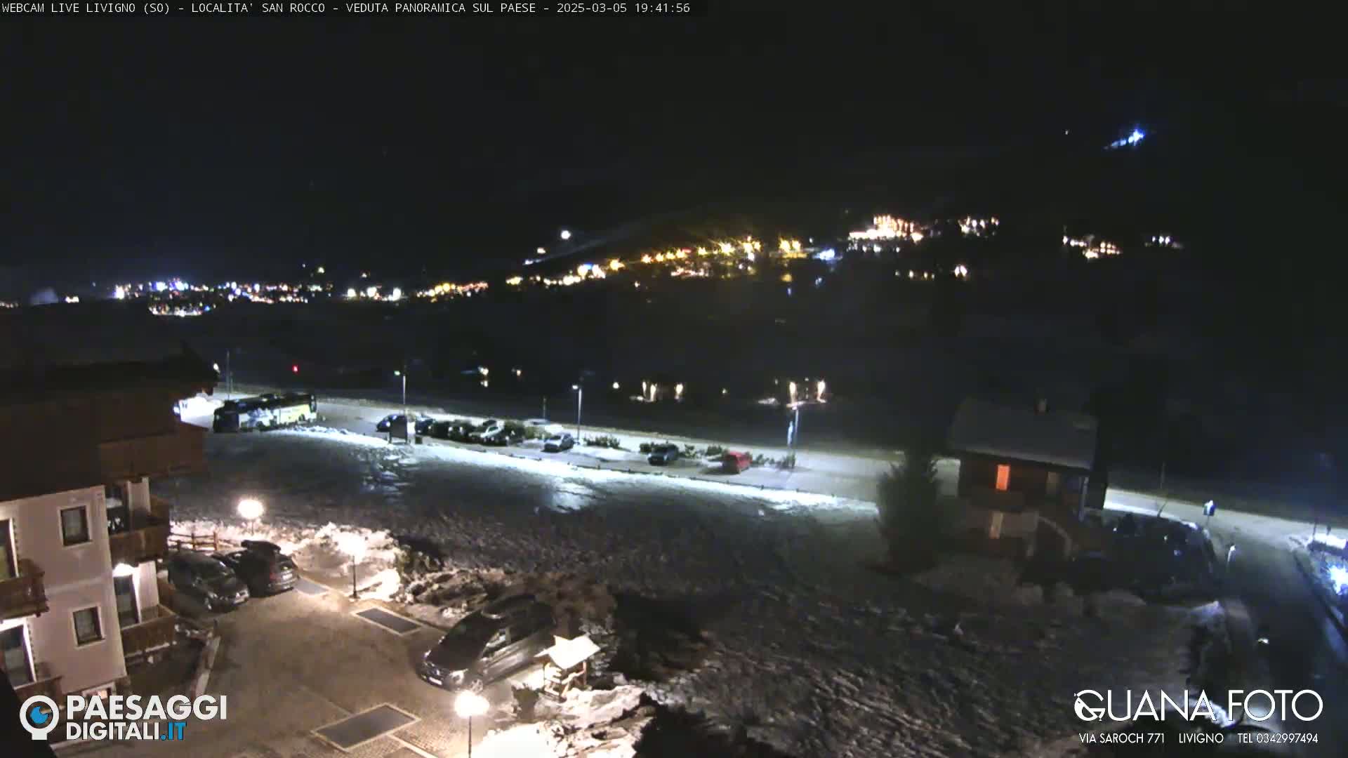 A nighttime, snow-covered parking lot and surrounding buildings are illuminated, with a distant town visible in the background under a clear, dark sky.