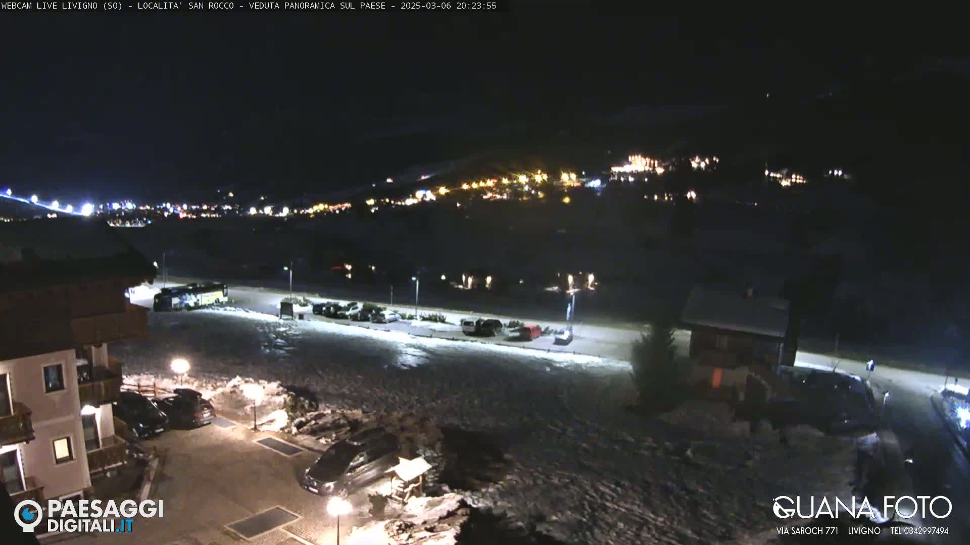 A nighttime, snow-covered parking lot and village scene with numerous lights visible in the distance.