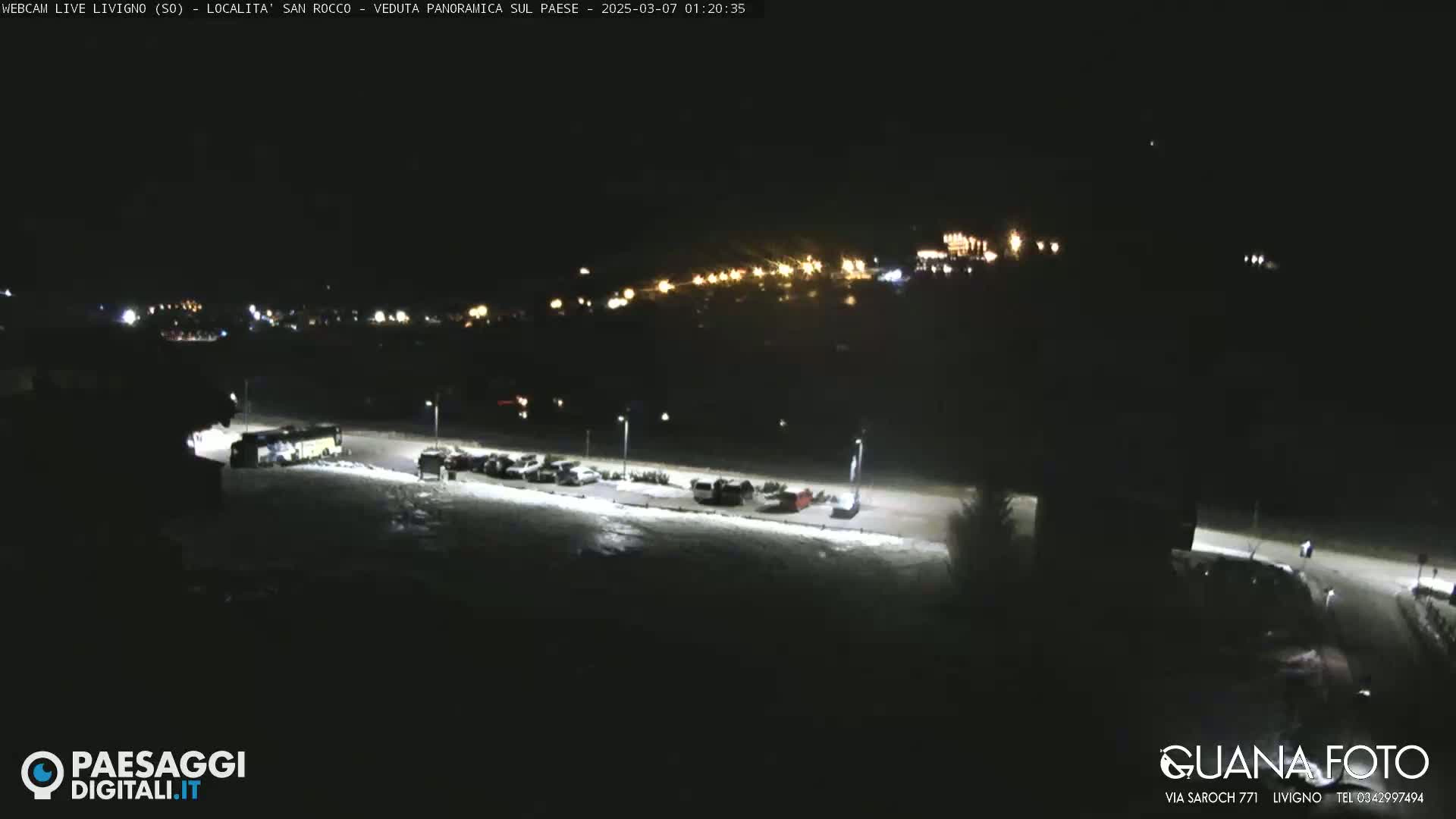 A snow-covered parking lot with several vehicles and a bus is illuminated at night, with a distant village showing faint lights in the background.
