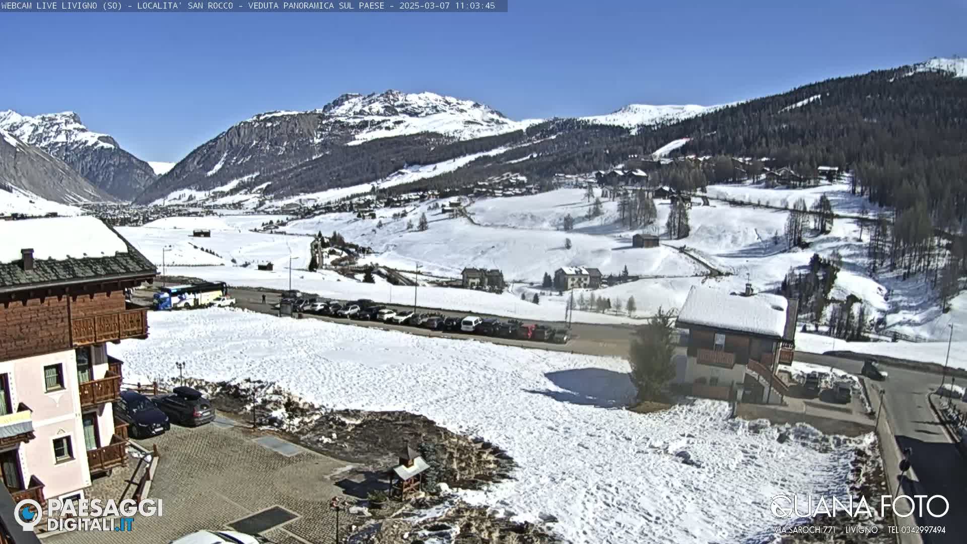 A sunny, snow-covered mountain village scene with cars parked in a lot, and buildings visible in the foreground and throughout the valley.