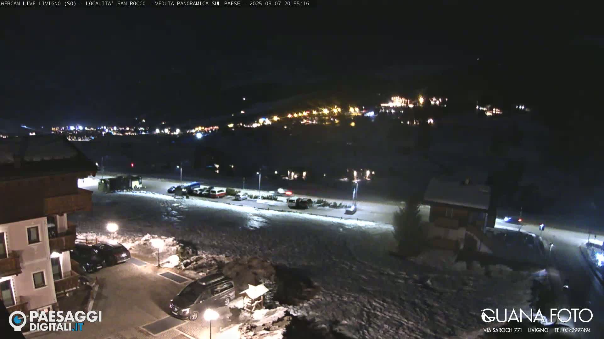 A nighttime, snow-covered parking lot and building complex overlook a small town illuminated by lights in the distance.