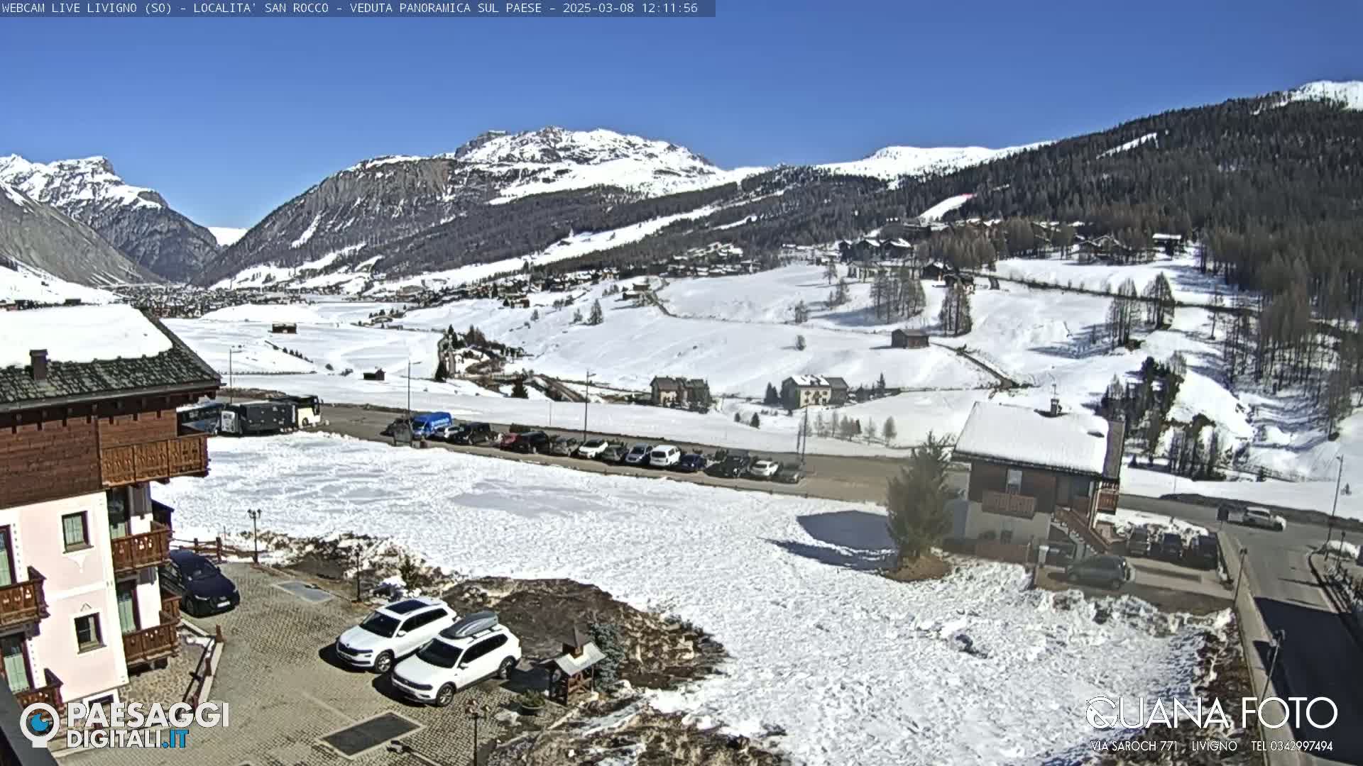 A sunny, snow-covered mountain valley with buildings, parked cars, and a few patches of bare ground.