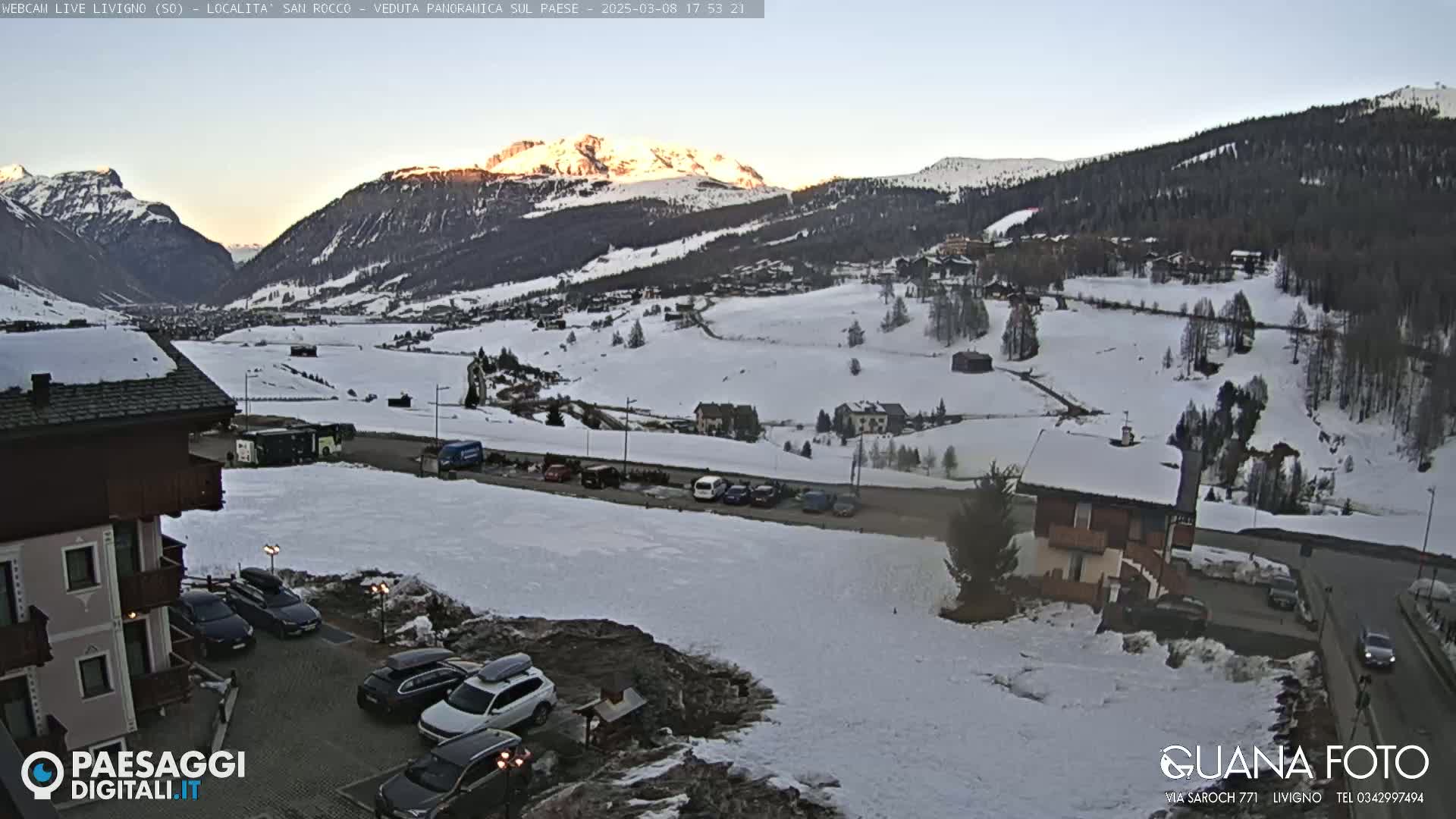 A snow-covered mountain valley at sunset, with a village and parking lot visible in the foreground.