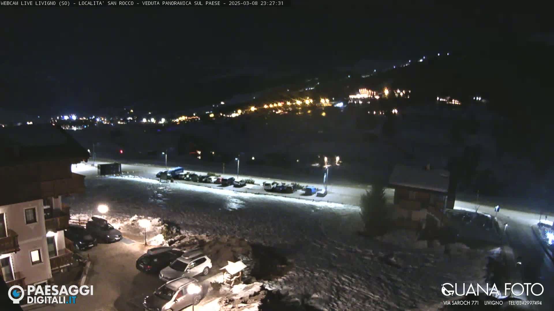 A nighttime, snow-covered parking lot and a village with illuminated buildings are visible under a dark sky.