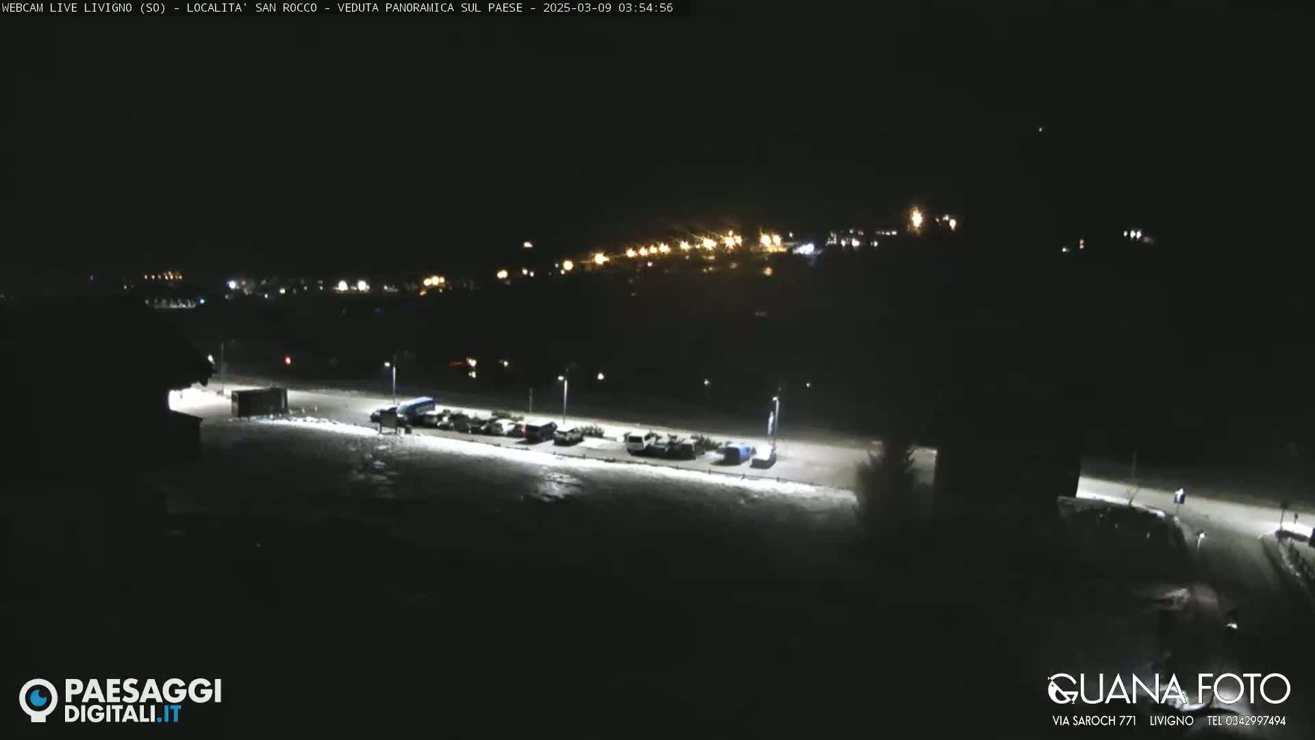 A nighttime, snow-covered parking lot with a few cars is visible, overlooking a dimly lit town in the distance.