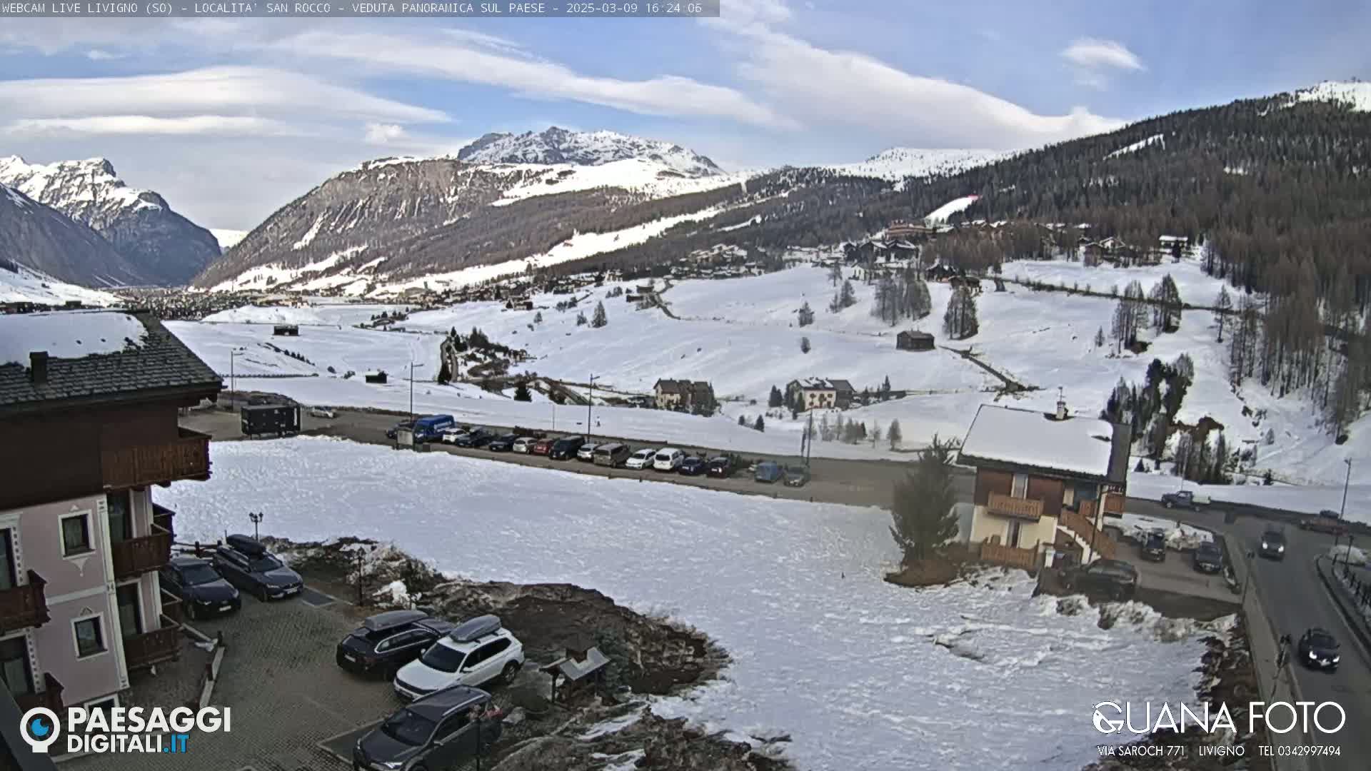 A snow-covered mountain valley village is viewed from a hillside, with cars parked in a lot near buildings and a partly sunny sky.