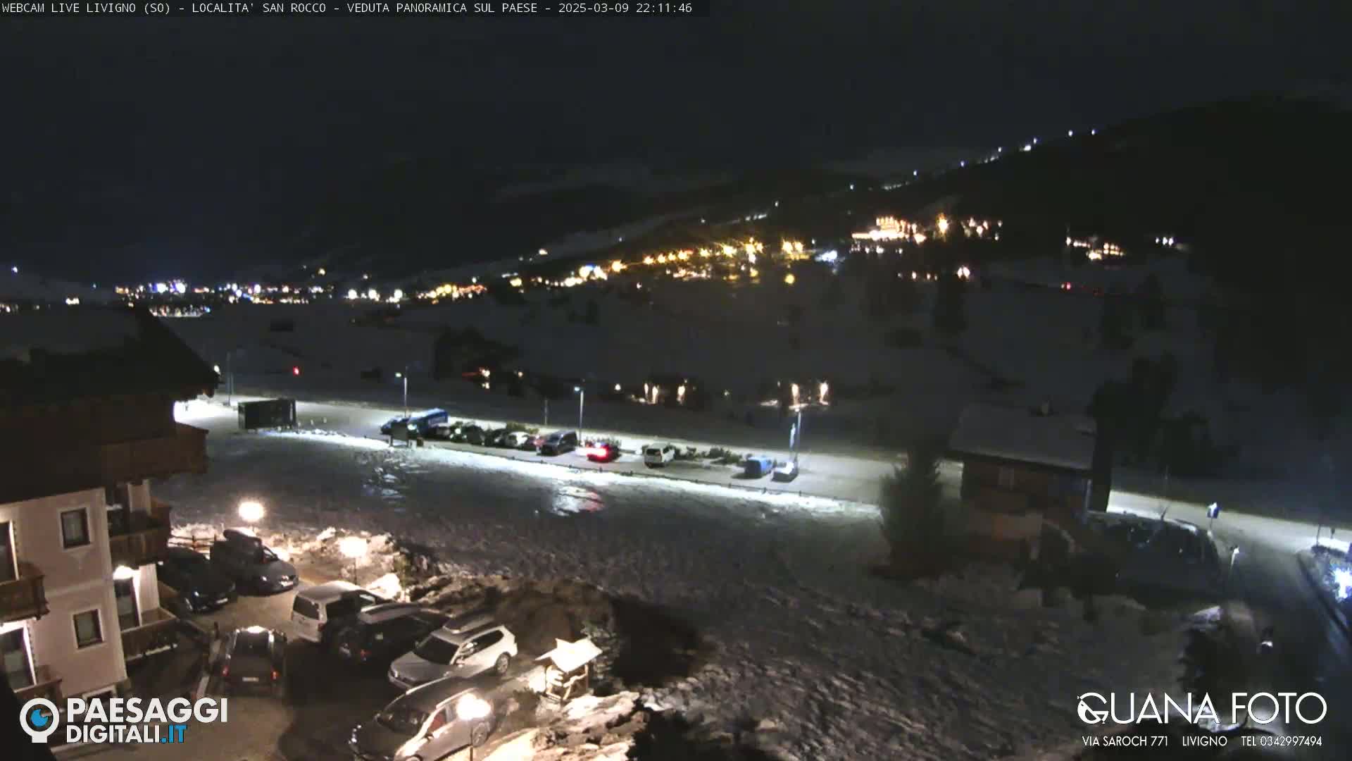 A nighttime, snow-covered parking lot and village are illuminated by artificial lights, with a hill in the background also showing some lights.