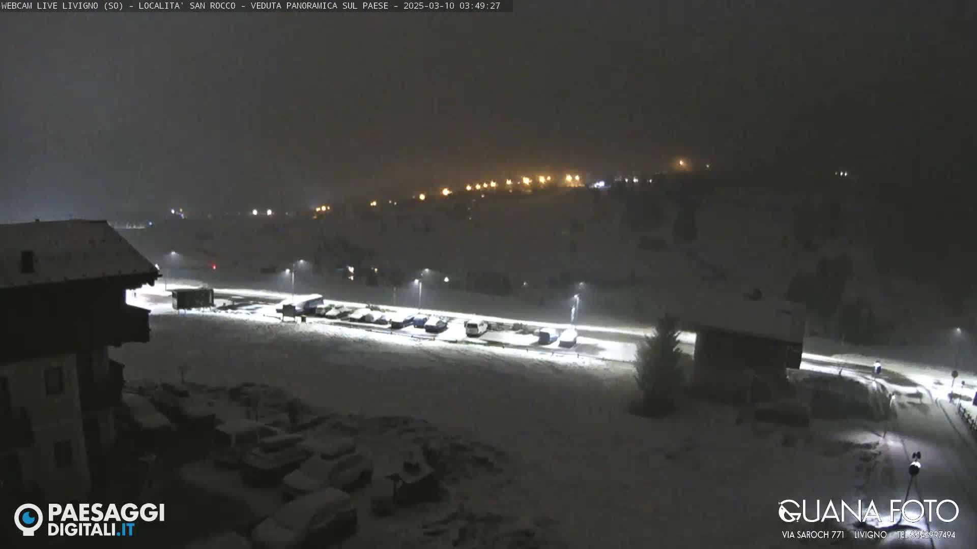 A nighttime snow-covered parking lot and buildings are illuminated by streetlights under a dark, overcast sky.
