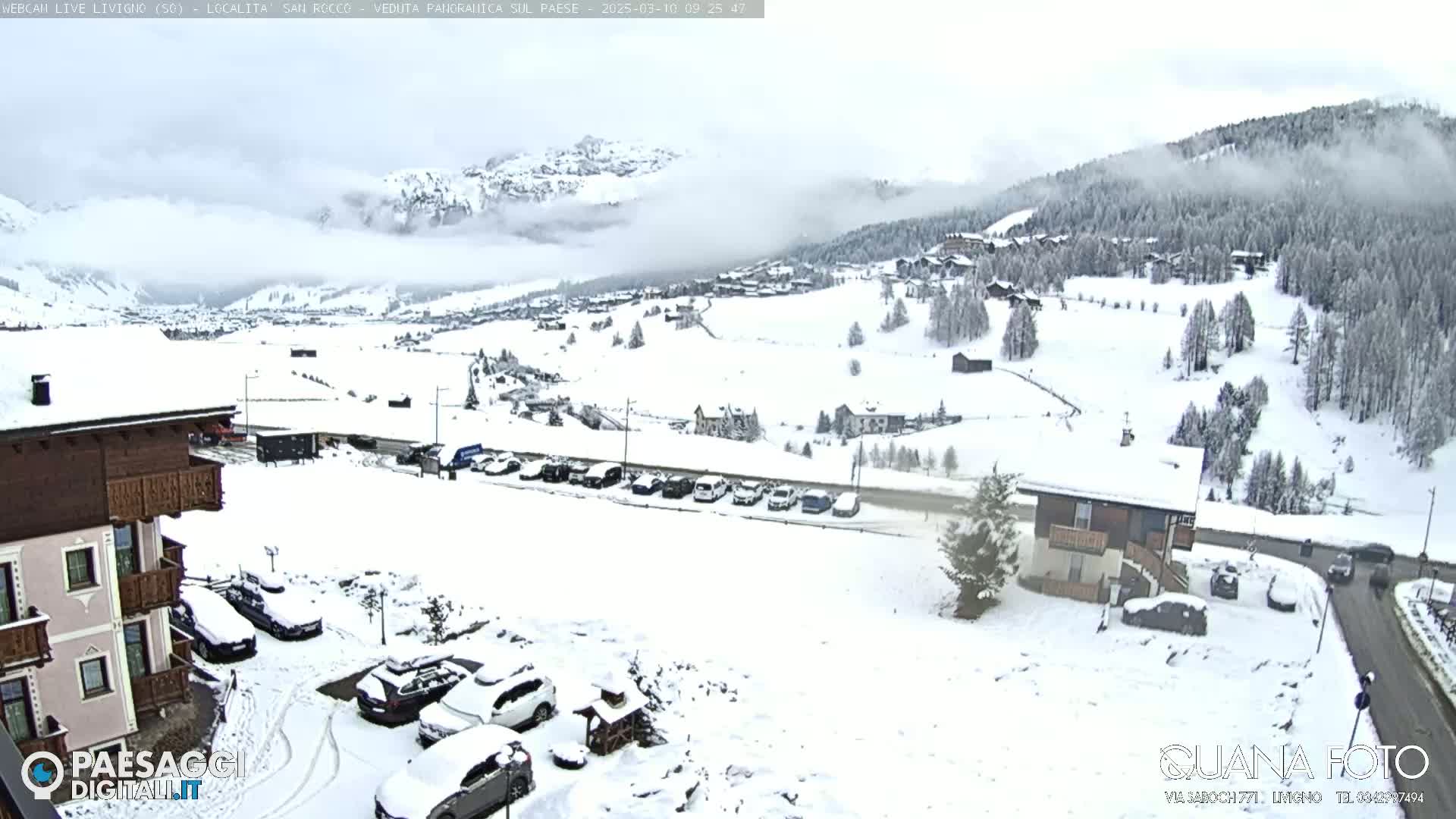 A snow-covered mountain village scene with overcast skies, featuring buildings, cars covered in snow, and a road winding through the landscape.