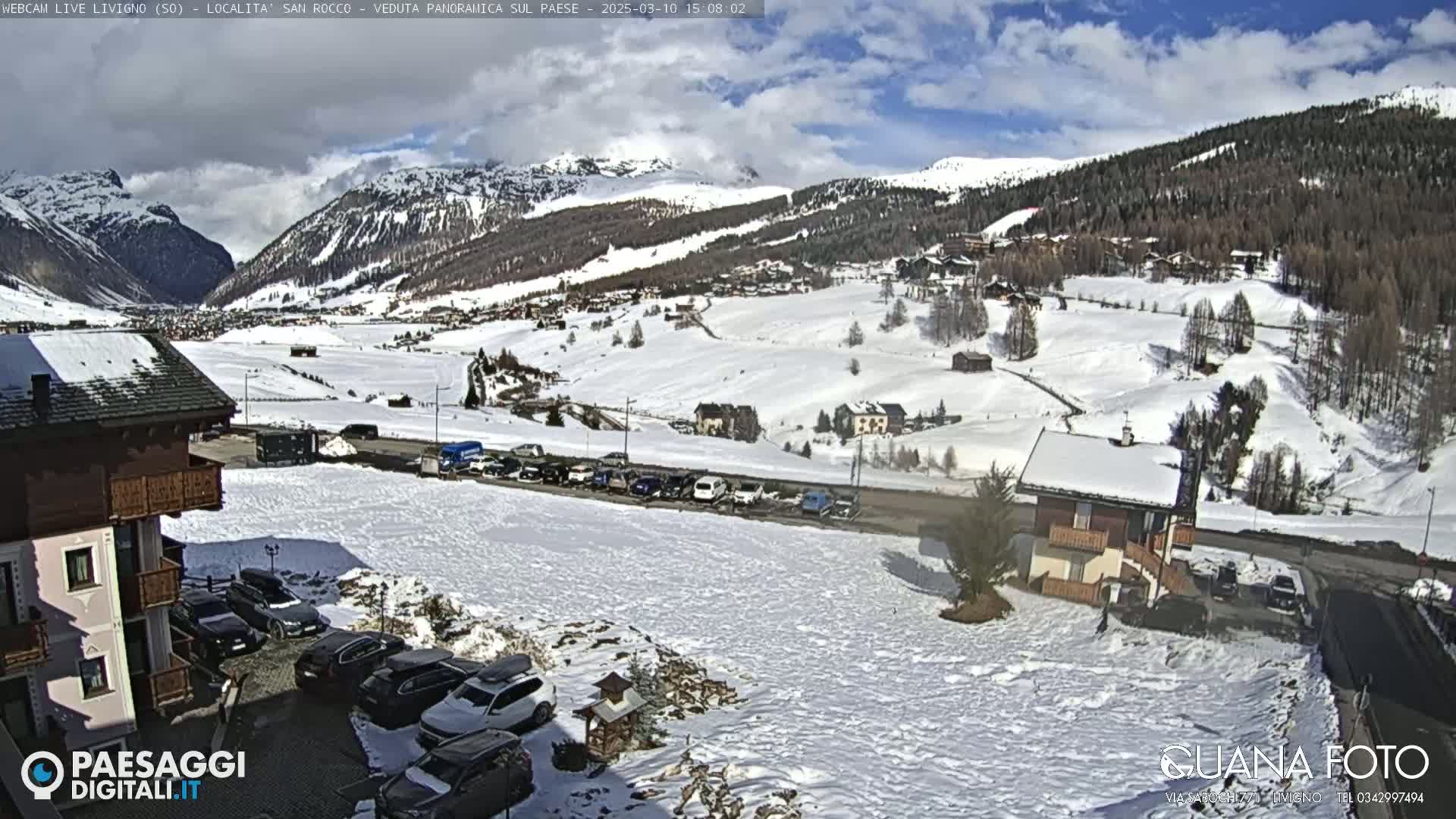 A snow-covered mountain valley town is viewed from a slightly elevated position on a partly cloudy day, with cars parked in a lot in the foreground.