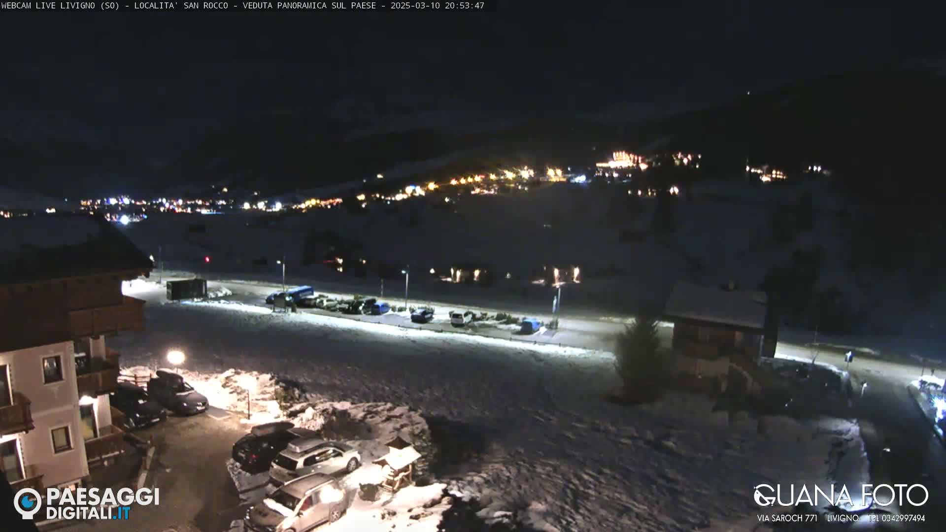 A nighttime, snow-covered parking lot and village are illuminated by streetlights and building lights, with mountains visible in the background under a clear, dark sky.