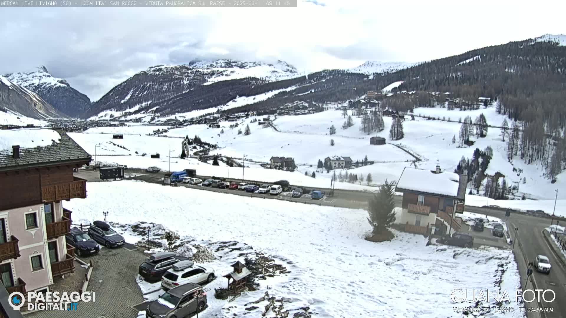 A snow-covered valley scene with several buildings, cars in parking lots, and mountains in the background under an overcast sky.
