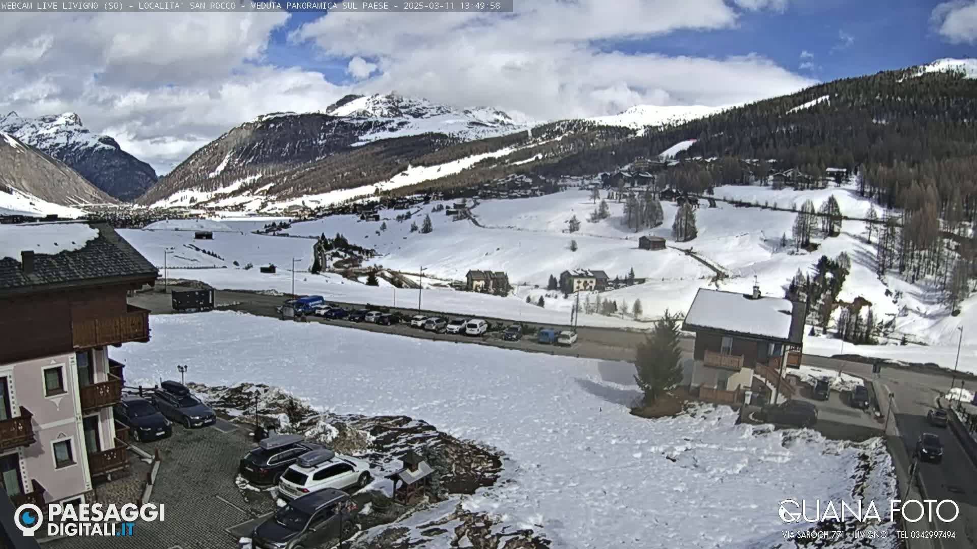 A snow-covered mountain valley with buildings and parked cars, under a partly cloudy sky.