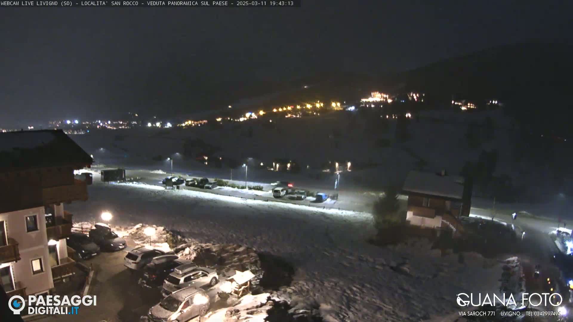 A nighttime, snow-covered scene shows a village lit up in the distance, with cars parked in a lot in the foreground near a building.