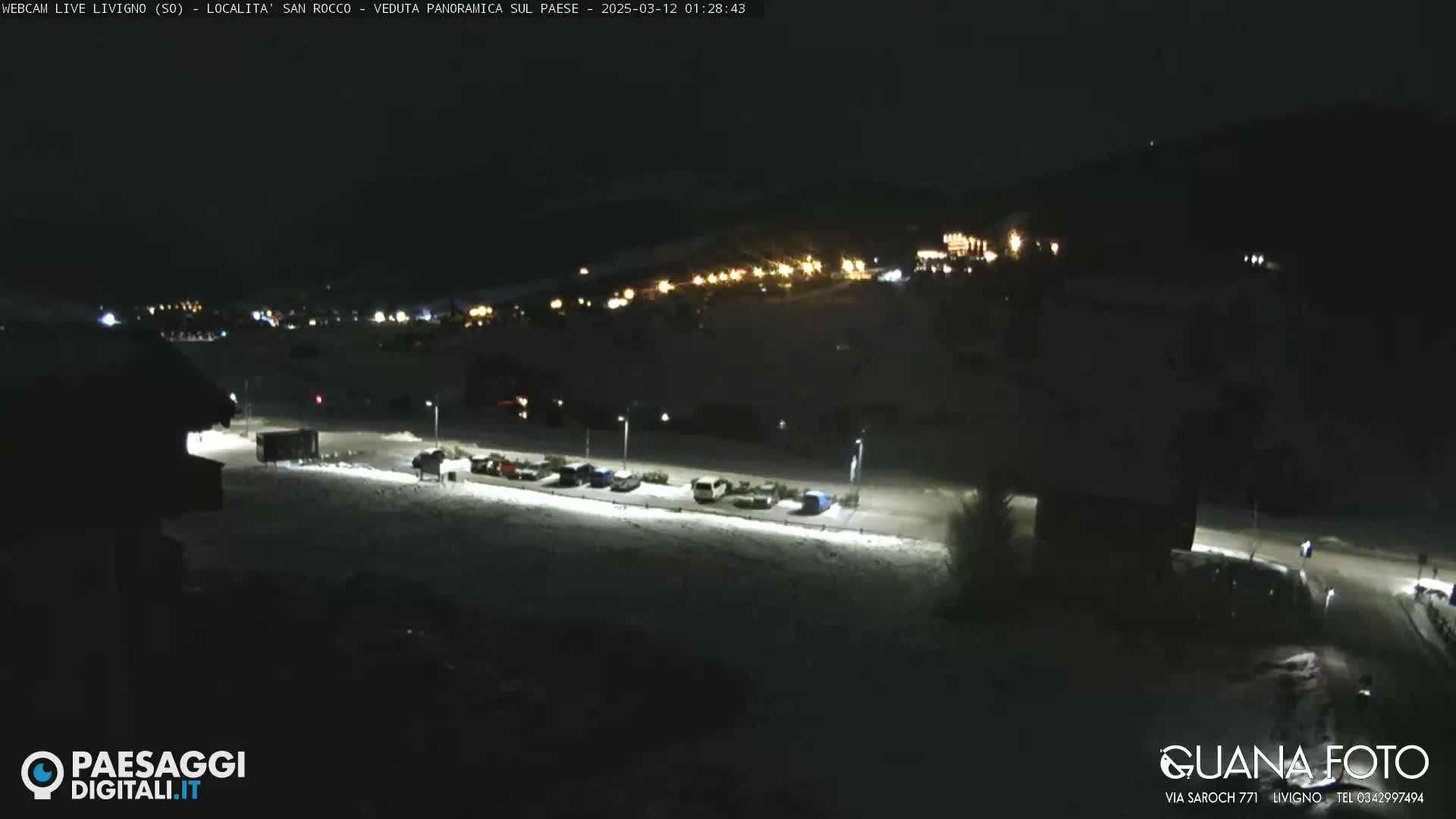 A nighttime, snow-covered parking lot with several cars and distant lights from buildings on a hill in the background.