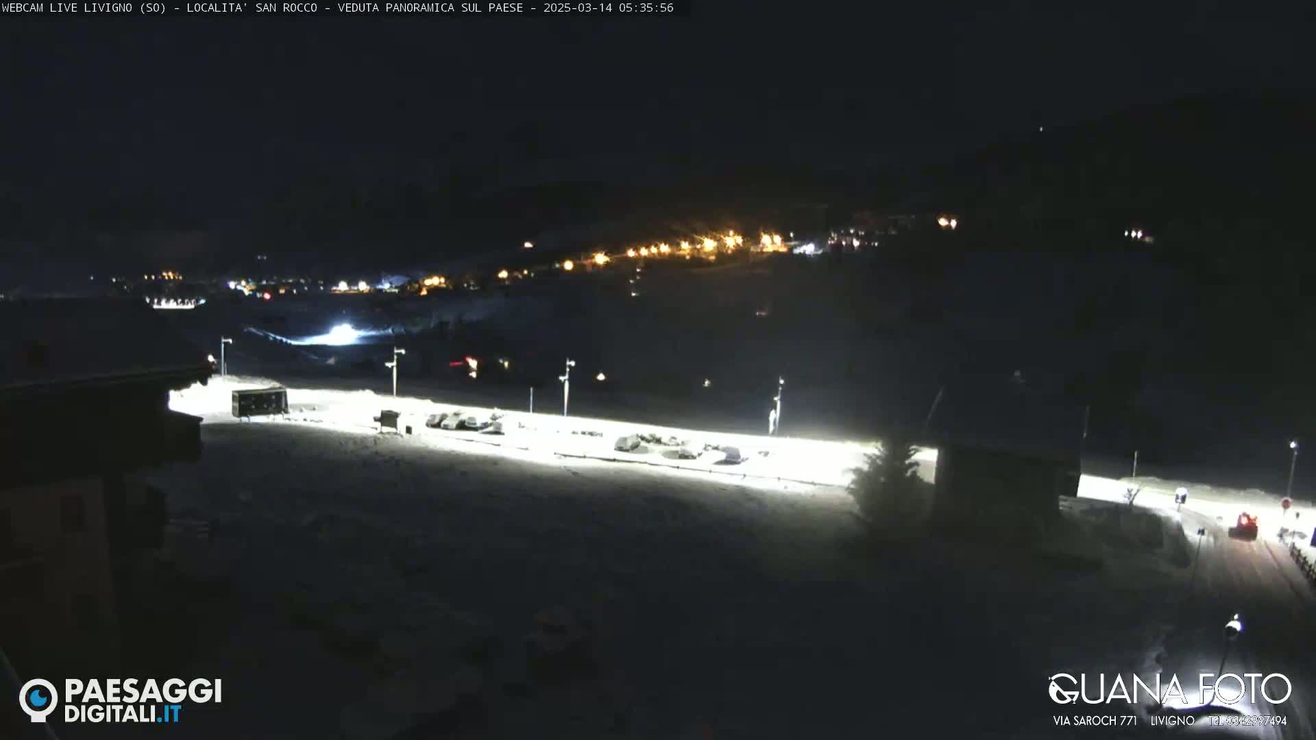 A snow-covered parking lot and surrounding area are illuminated at night, with a town's lights visible in the distance.
