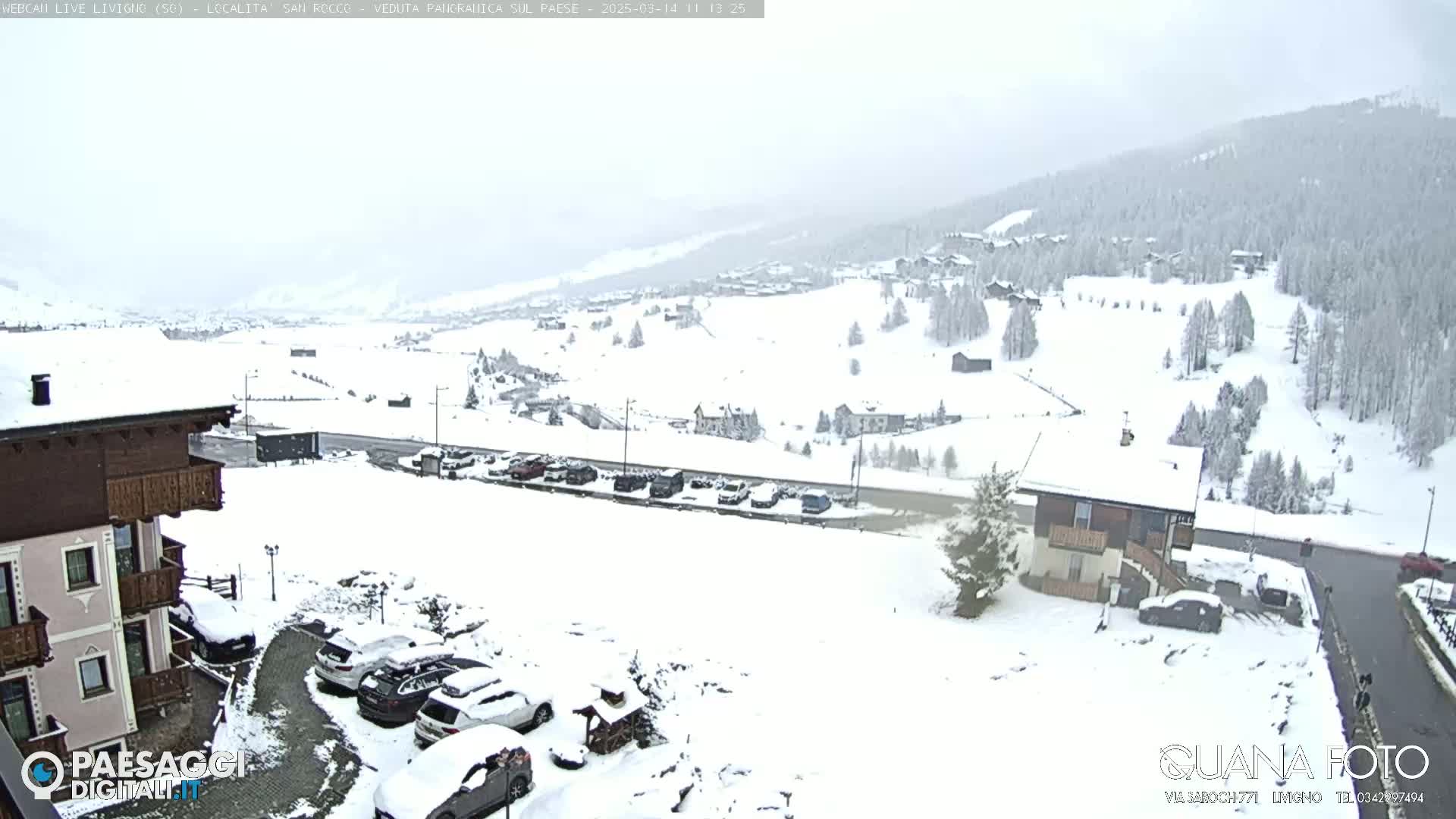A snow-covered village nestled in a mountainous region, viewed from a slightly elevated position, showing a parking lot with several cars, and buildings partially obscured by snow.