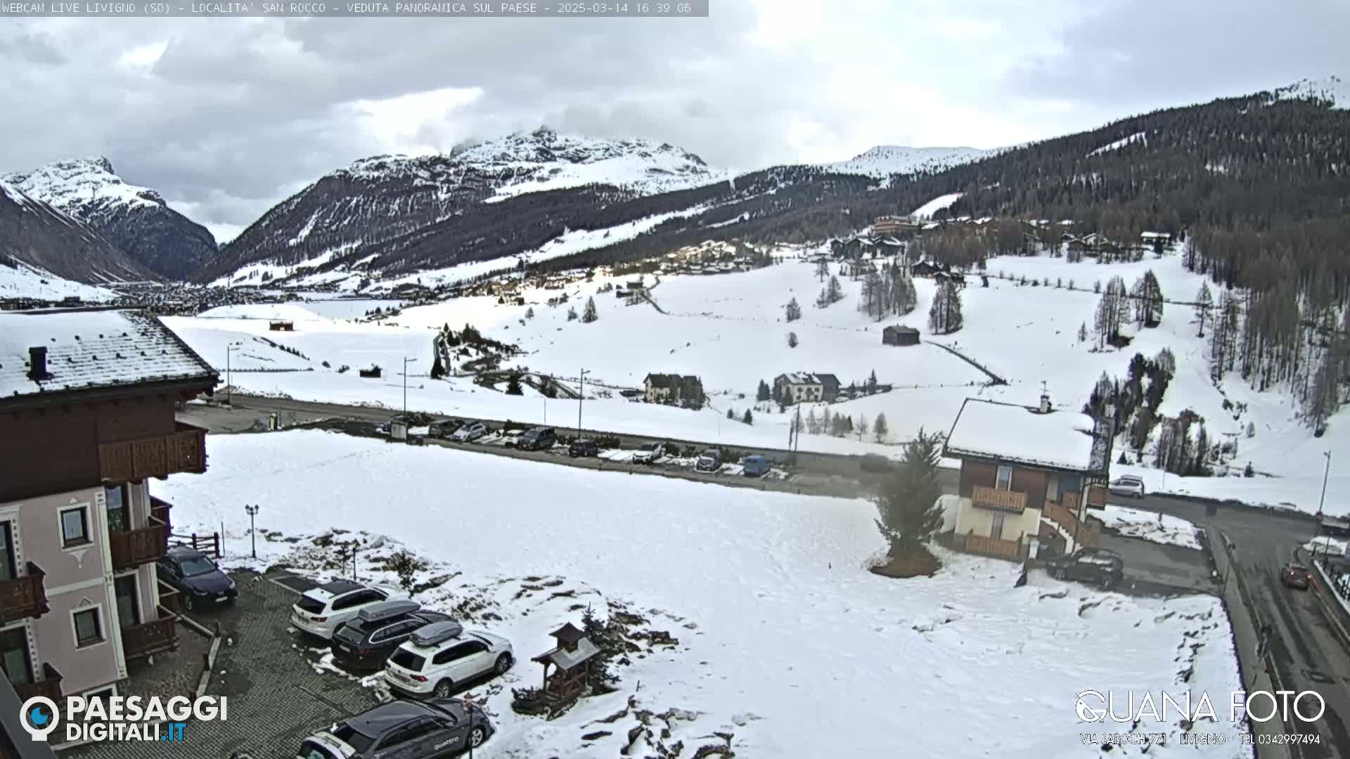 A snow-covered mountain valley scene with overcast skies, featuring a residential area with parked cars and buildings, viewed from an elevated vantage point.