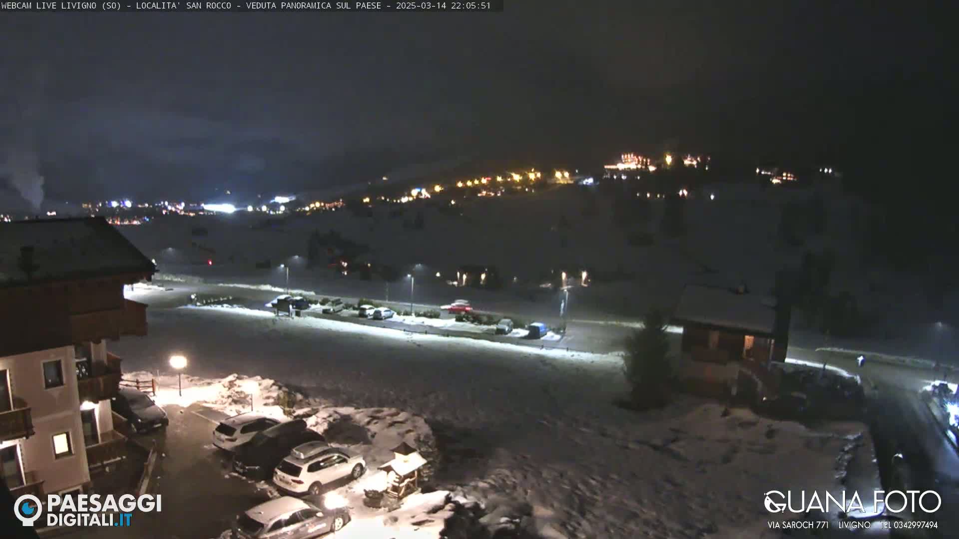 A nighttime, snow-covered landscape shows a village with illuminated buildings in the distance, a parking lot with several cars, and a building in the foreground under a dark sky.