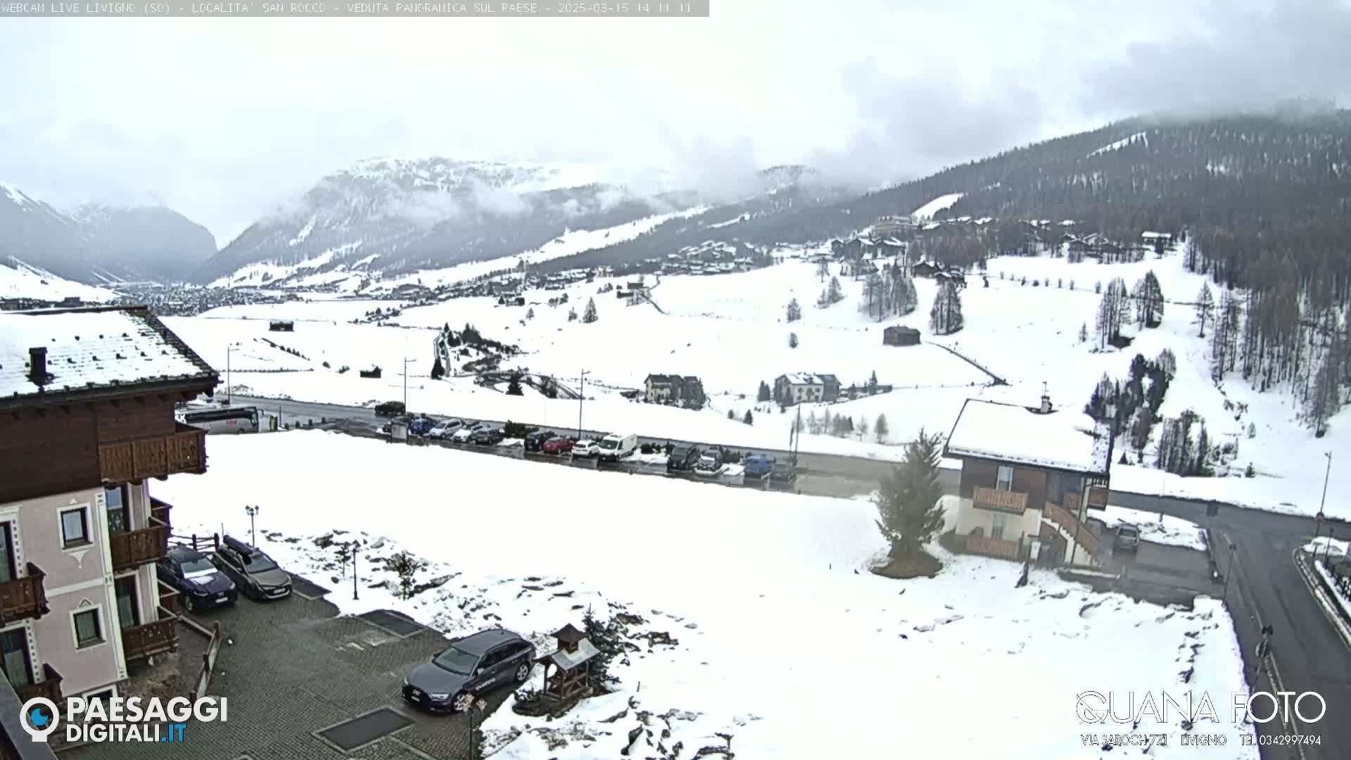 A snow-covered mountain valley scene with buildings, cars parked in a lot, and overcast skies.