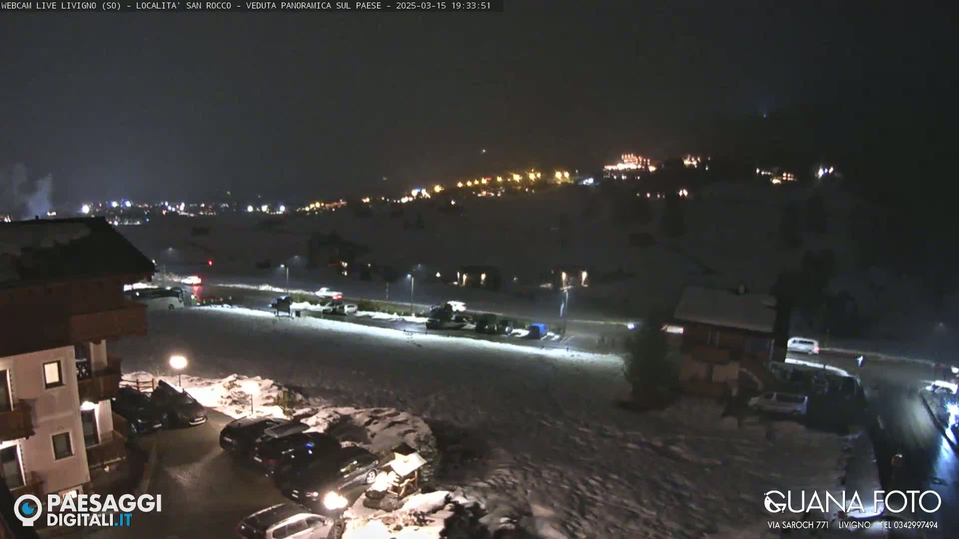 A nighttime, snow-covered parking lot and residential area with a few cars and buildings, illuminated by streetlights, overlooking a town speckled with distant lights.