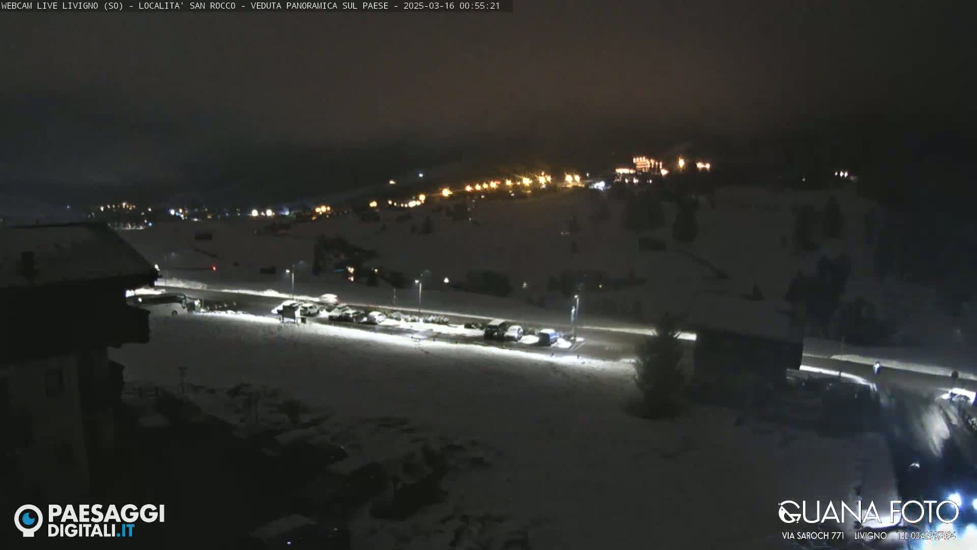 A nighttime, snow-covered scene shows a village with sparsely parked cars illuminated by streetlights.