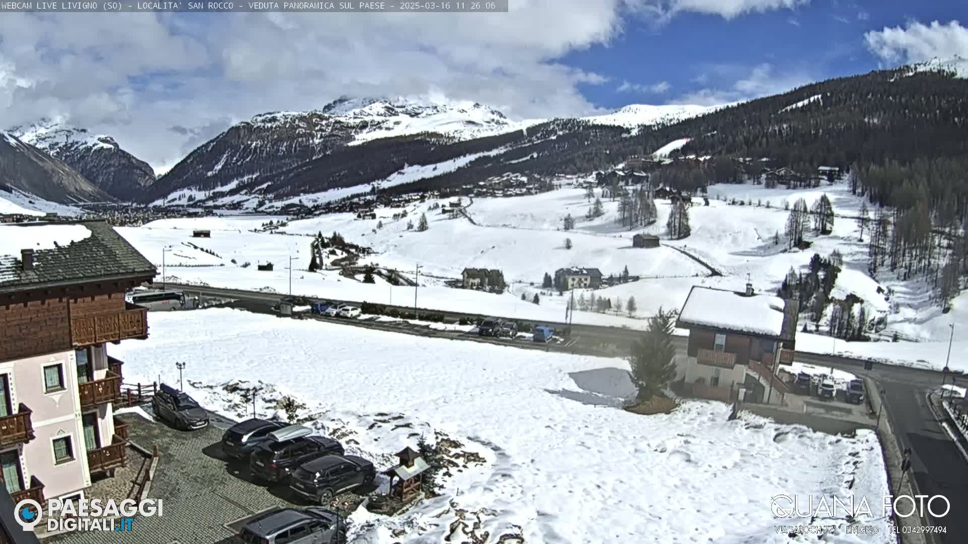 A snow-covered valley nestled between mountains under a partly cloudy sky, with cars parked near buildings in the foreground.