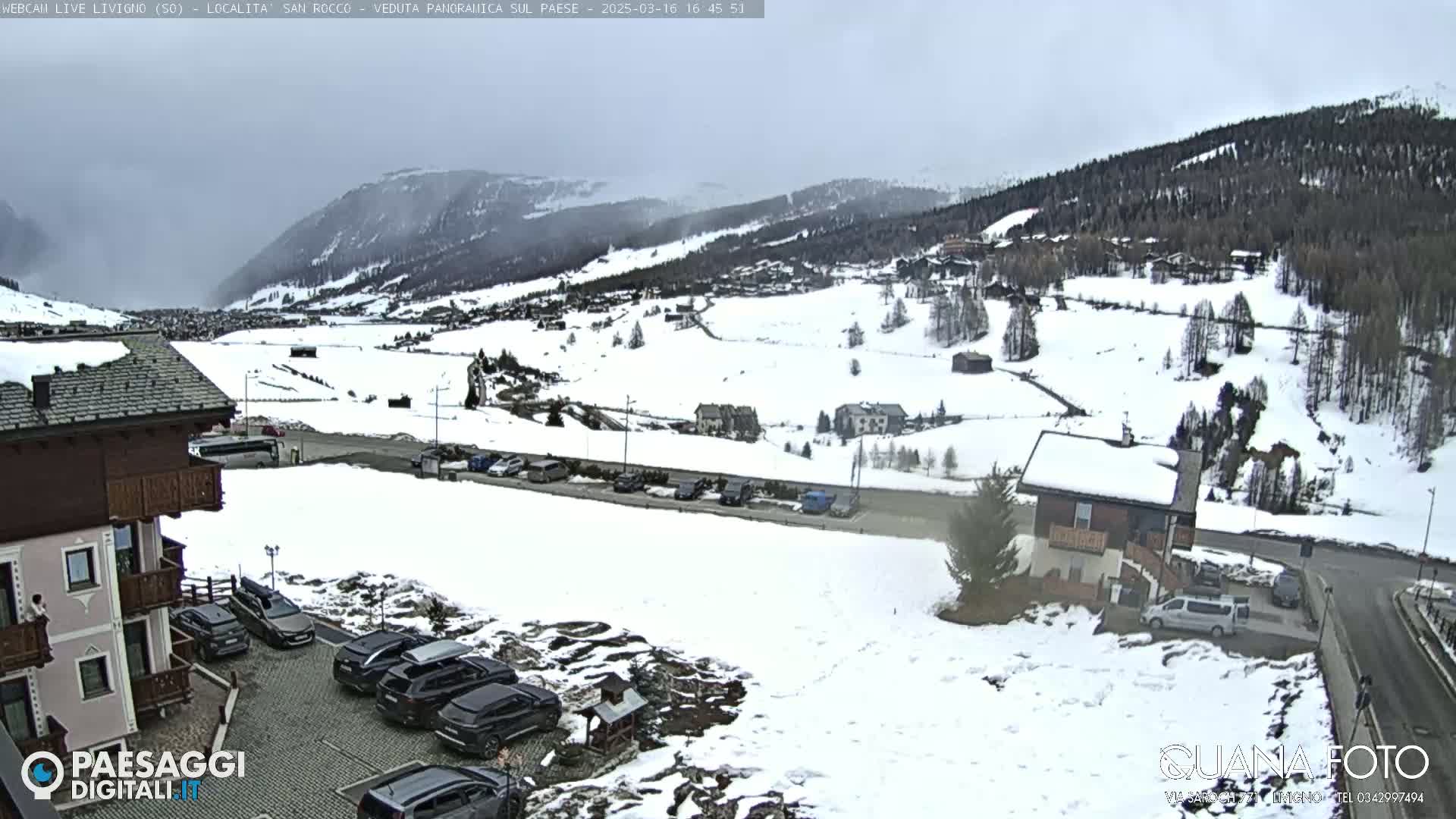 A snow-covered mountain valley with buildings, parking lots, and vehicles under an overcast sky.