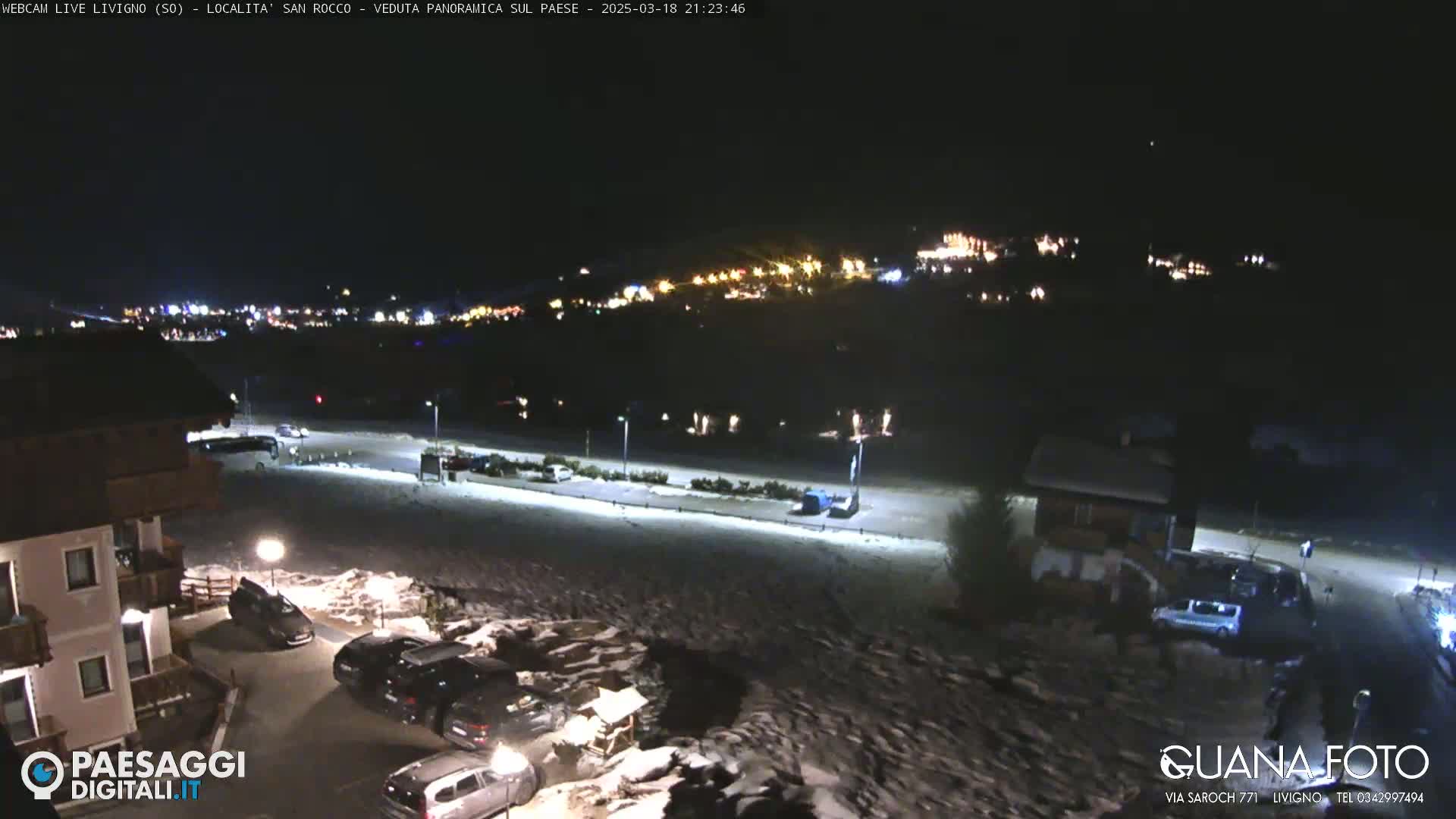 A nighttime, snow-covered parking lot and roadway are shown, with a village's lights visible in the distance.