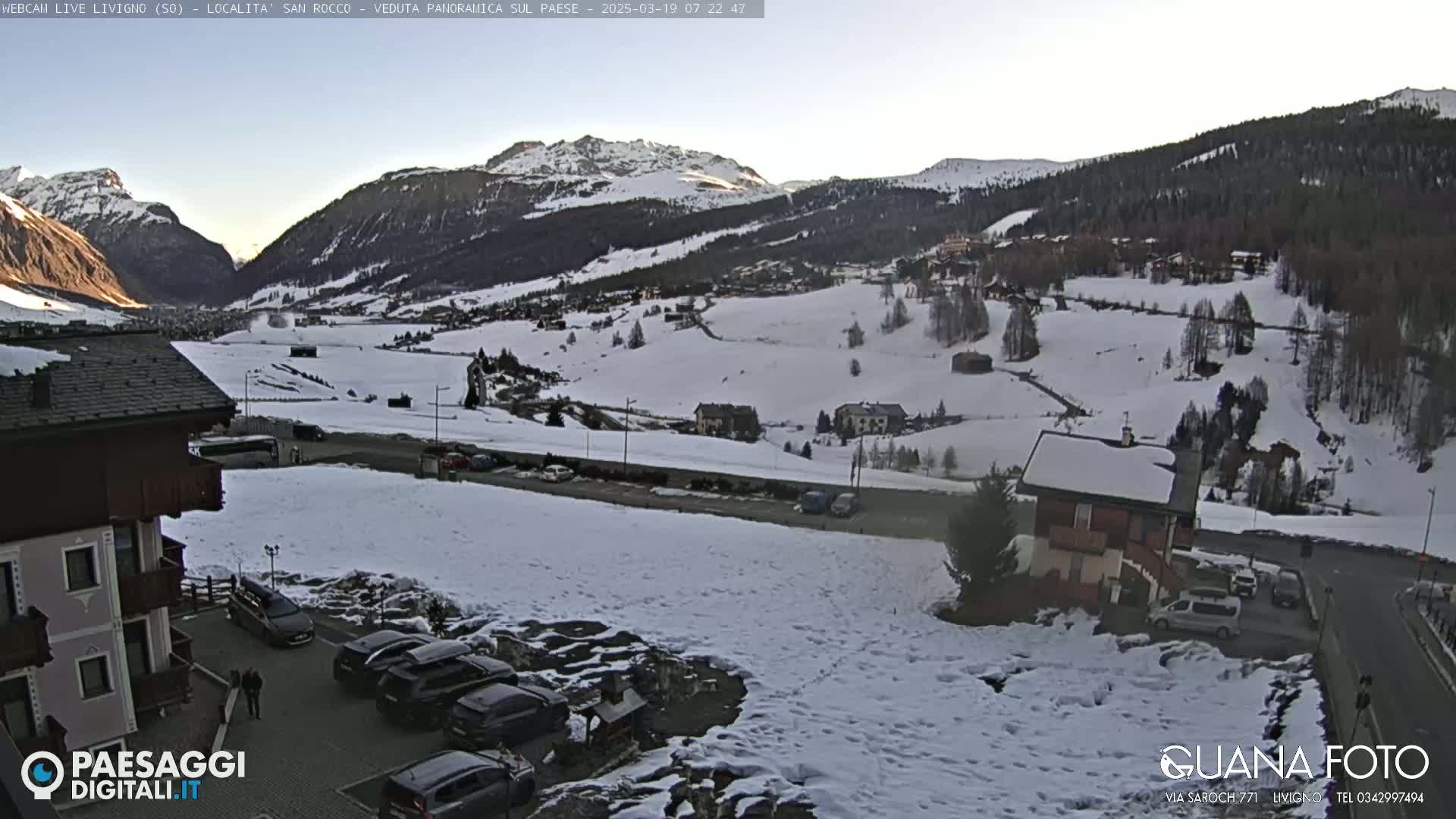 A snow-covered mountain valley village is viewed from a high vantage point, with several cars parked near buildings and a road visible in the foreground.