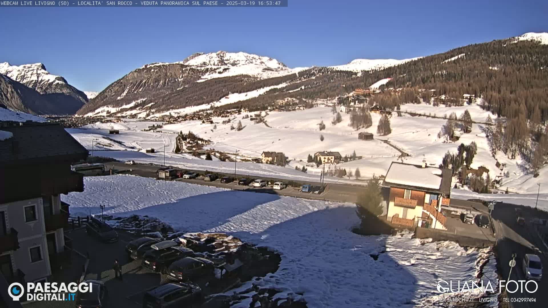 A sunny, snow-covered mountain valley scene with buildings, cars, and patches of bare ground, viewed from an elevated position.