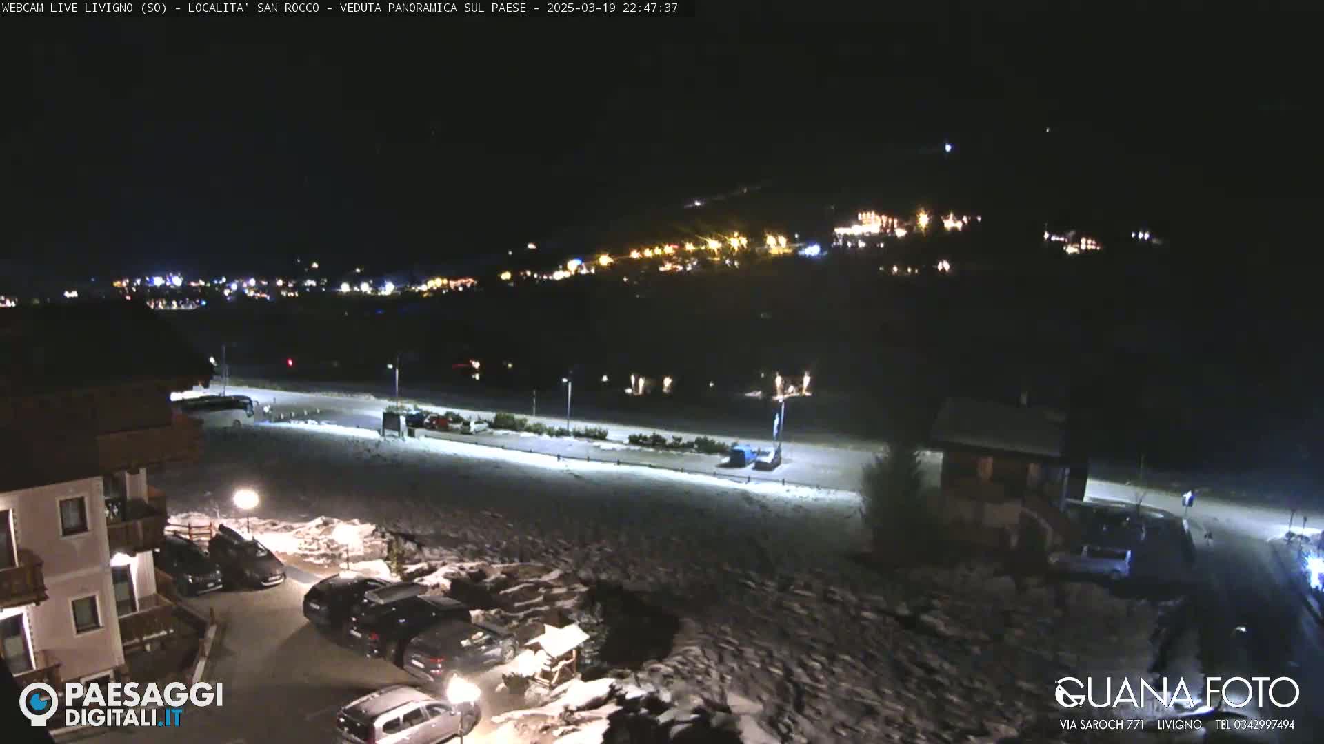 A nighttime view of a snow-covered parking lot and town with distant lights, under a clear sky.