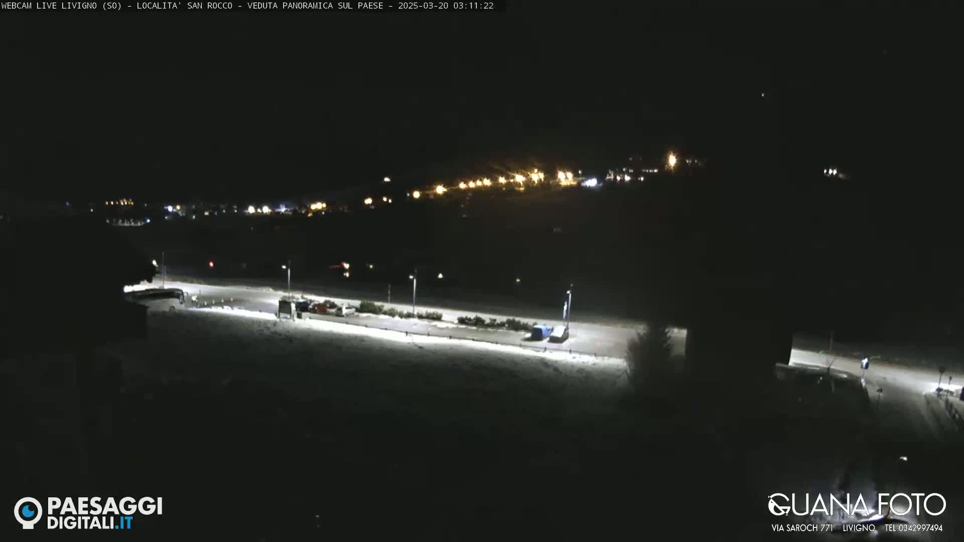 A nighttime, snow-covered parking lot and road are illuminated by streetlights, with a distant town faintly visible in the background.