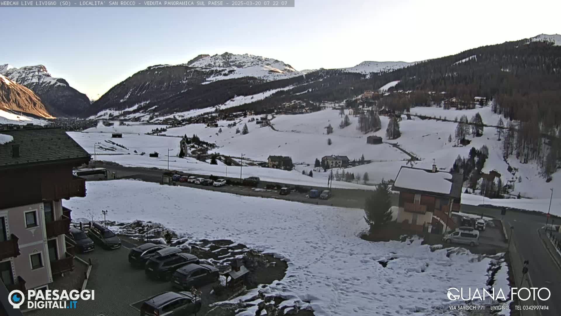A snow-covered mountain valley scene with several buildings, cars parked in lots, and a clear sky.