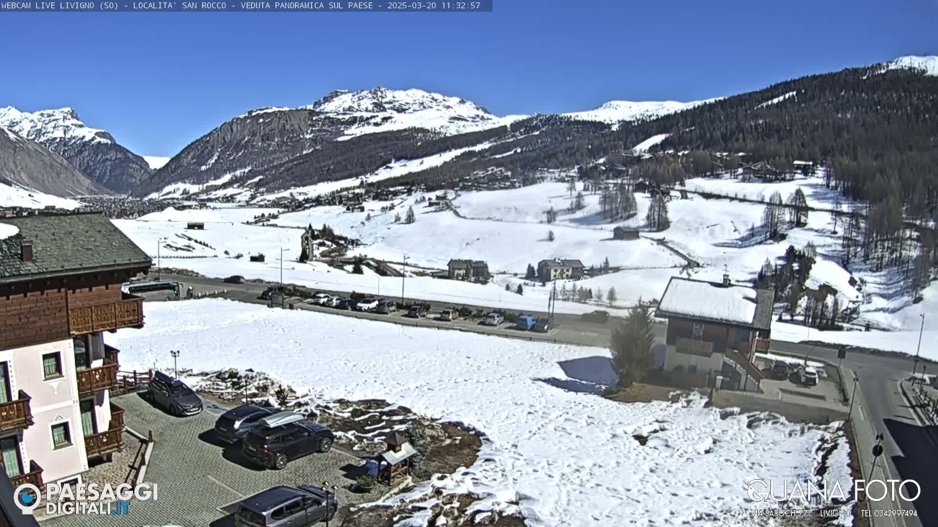 A sunny, snow-covered mountain valley scene with buildings and parked cars, viewed from a slightly elevated position.