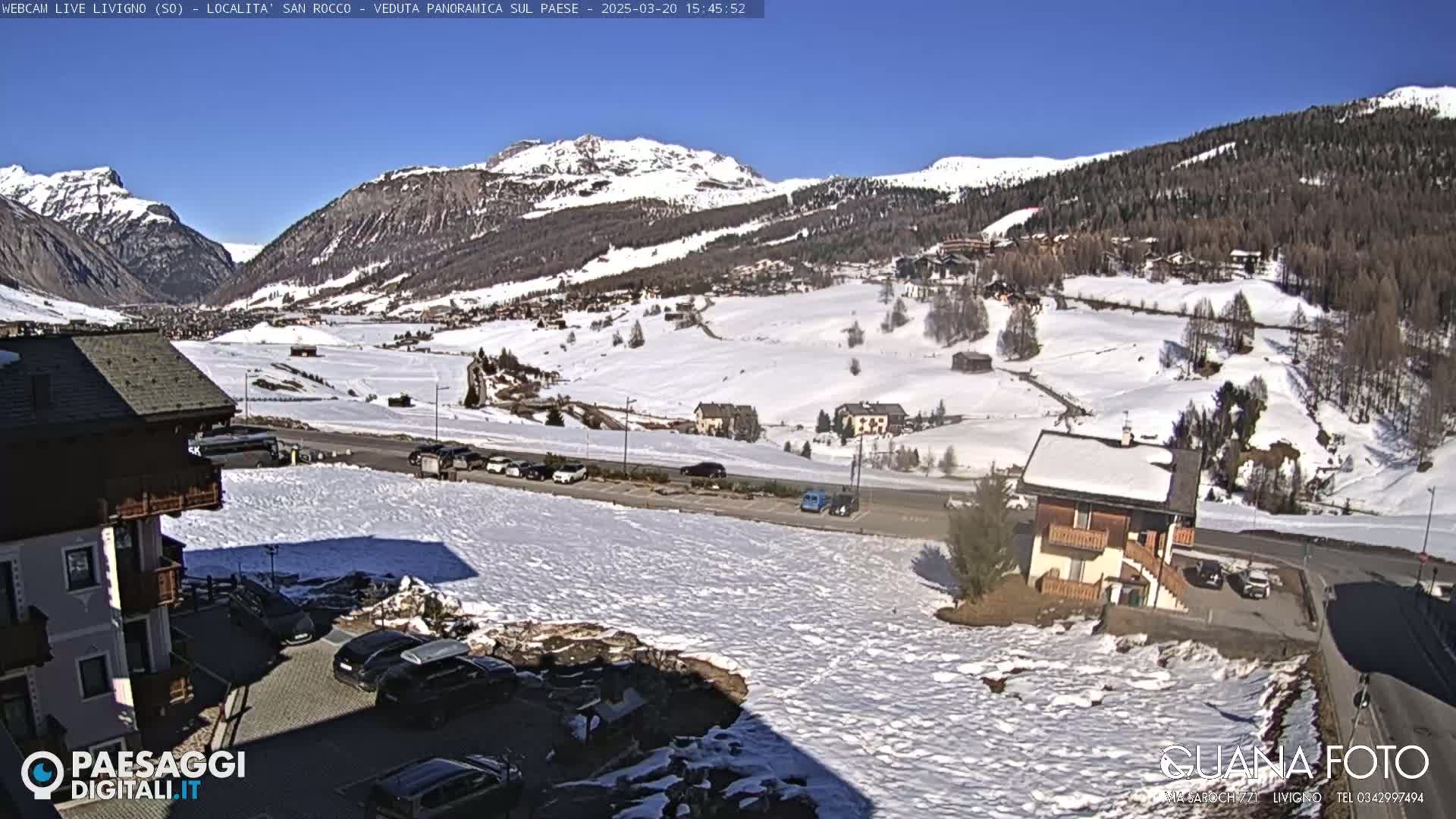 A sunny, snow-covered mountain valley scene with several buildings and cars visible, some of which are parked.