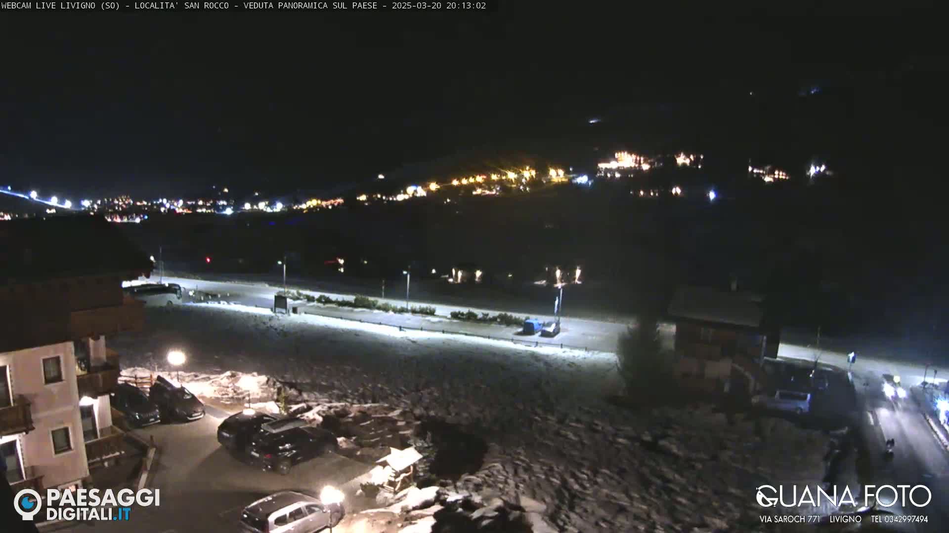 A nighttime, snow-covered parking lot and road are visible, with a village's lights illuminating the hillside in the distance.