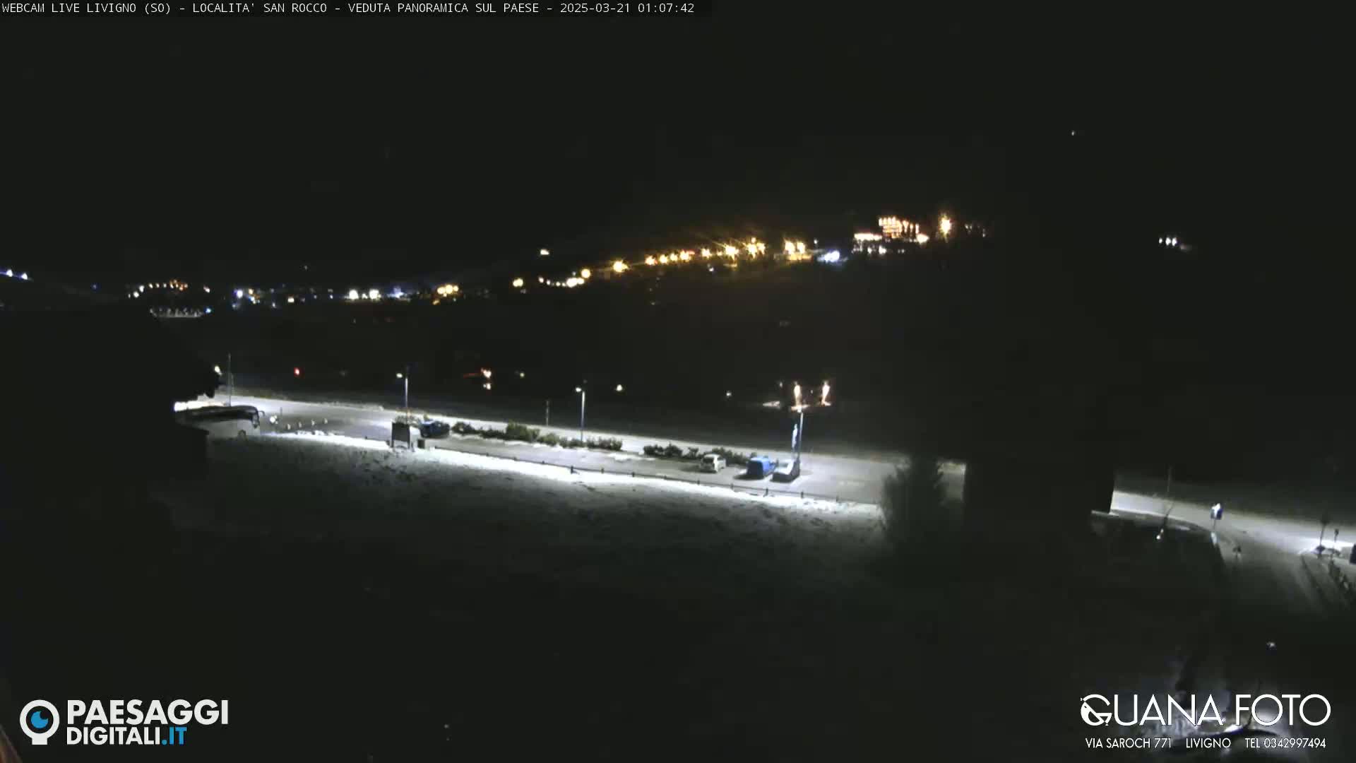 A nighttime, snow-covered parking lot and road are illuminated by streetlights, with a distant village glowing faintly in the background.