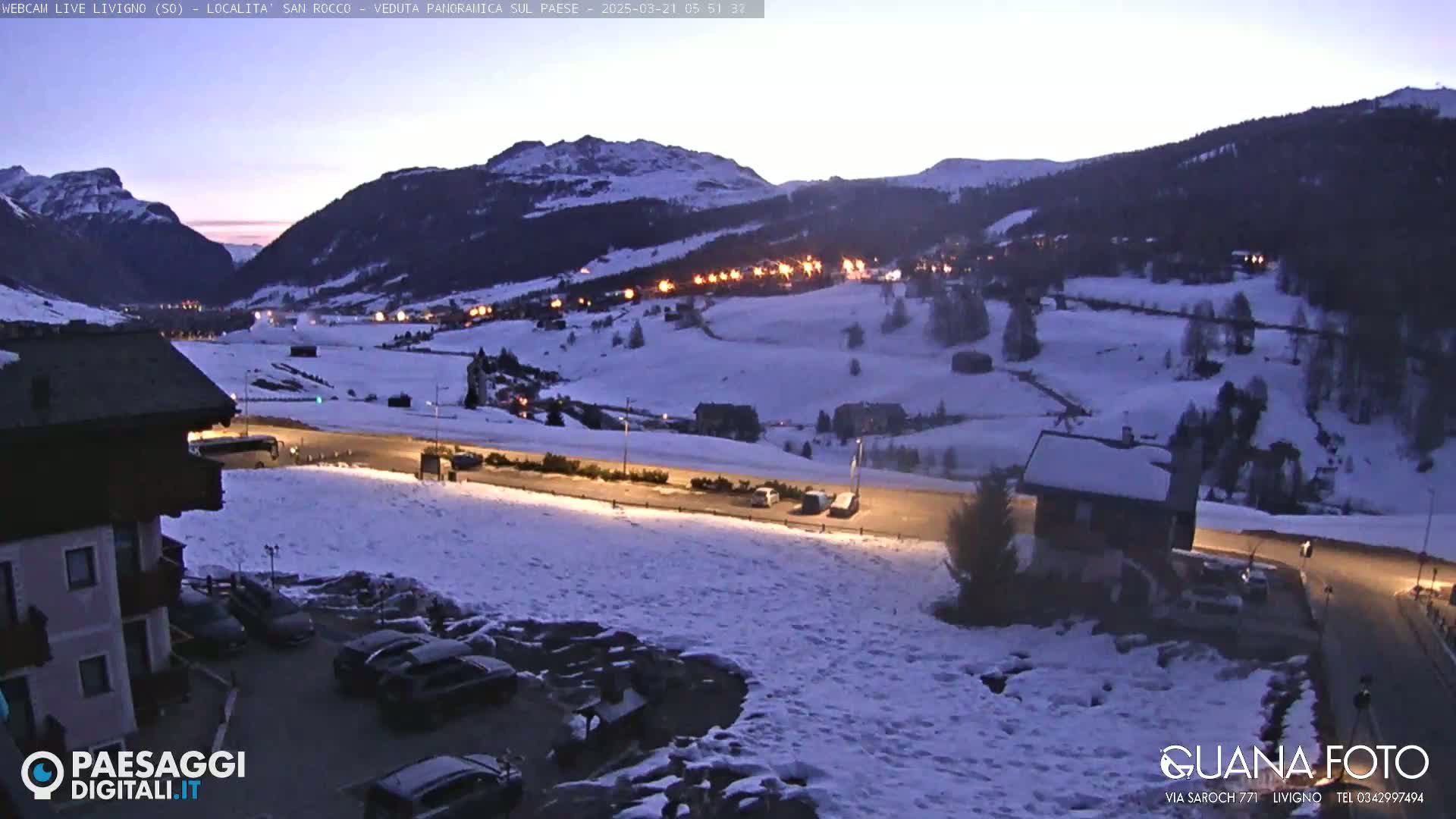 A snow-covered mountain village at twilight, with some lights on in the buildings and cars parked in a snowy lot.