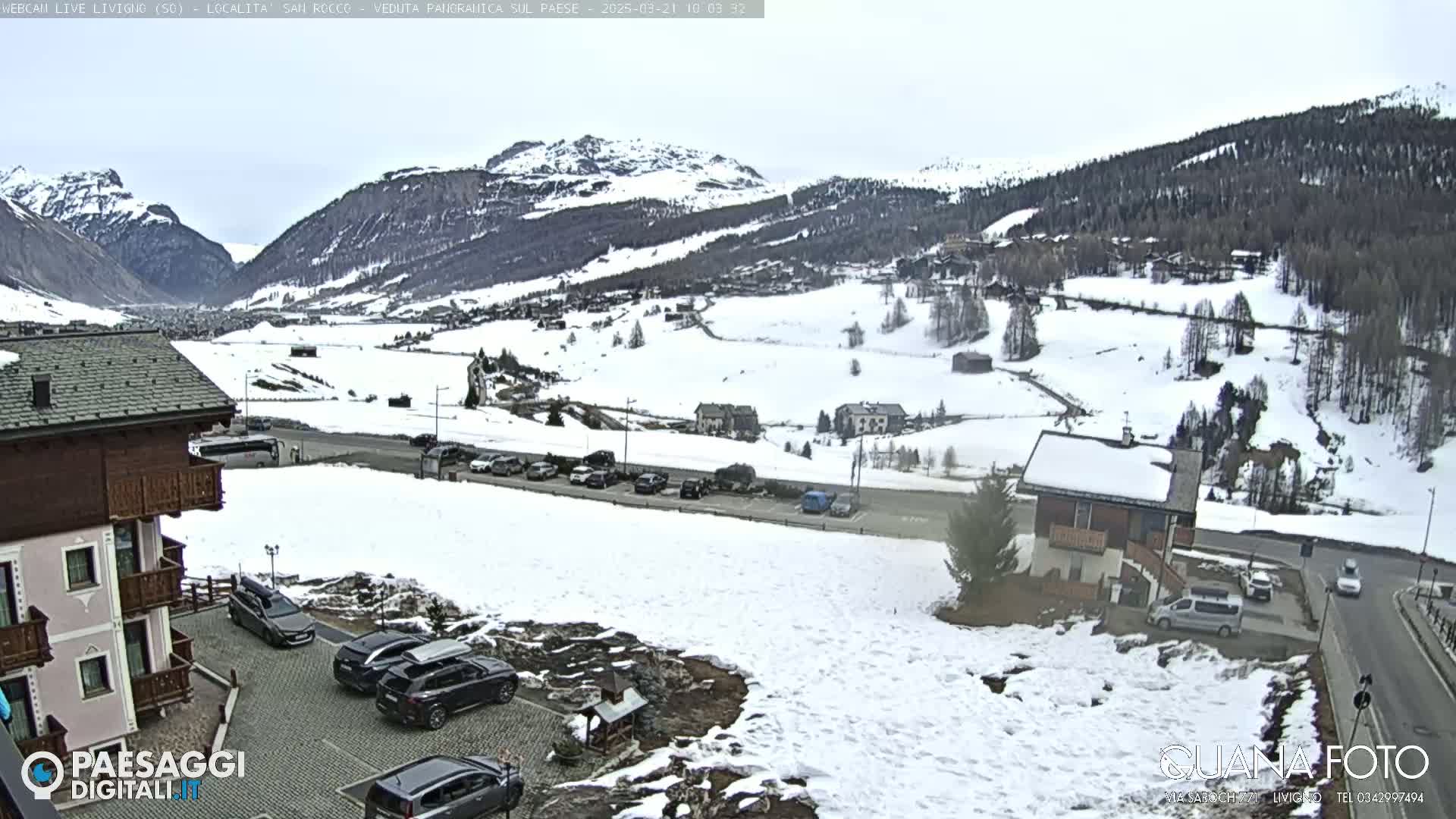 A snow-covered mountain valley scene is visible from a high vantage point, showing several buildings, cars parked in lots, and a road winding through the landscape.