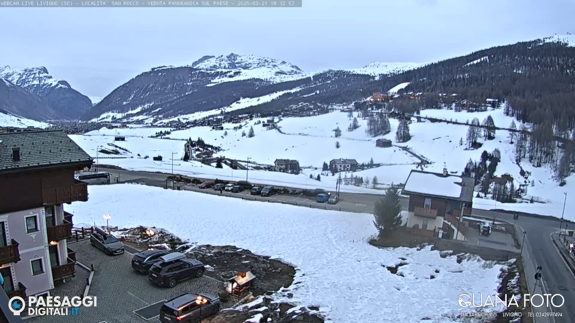 A snow-covered mountain valley scene with several buildings and parked cars, under a cloudy, twilight sky.
