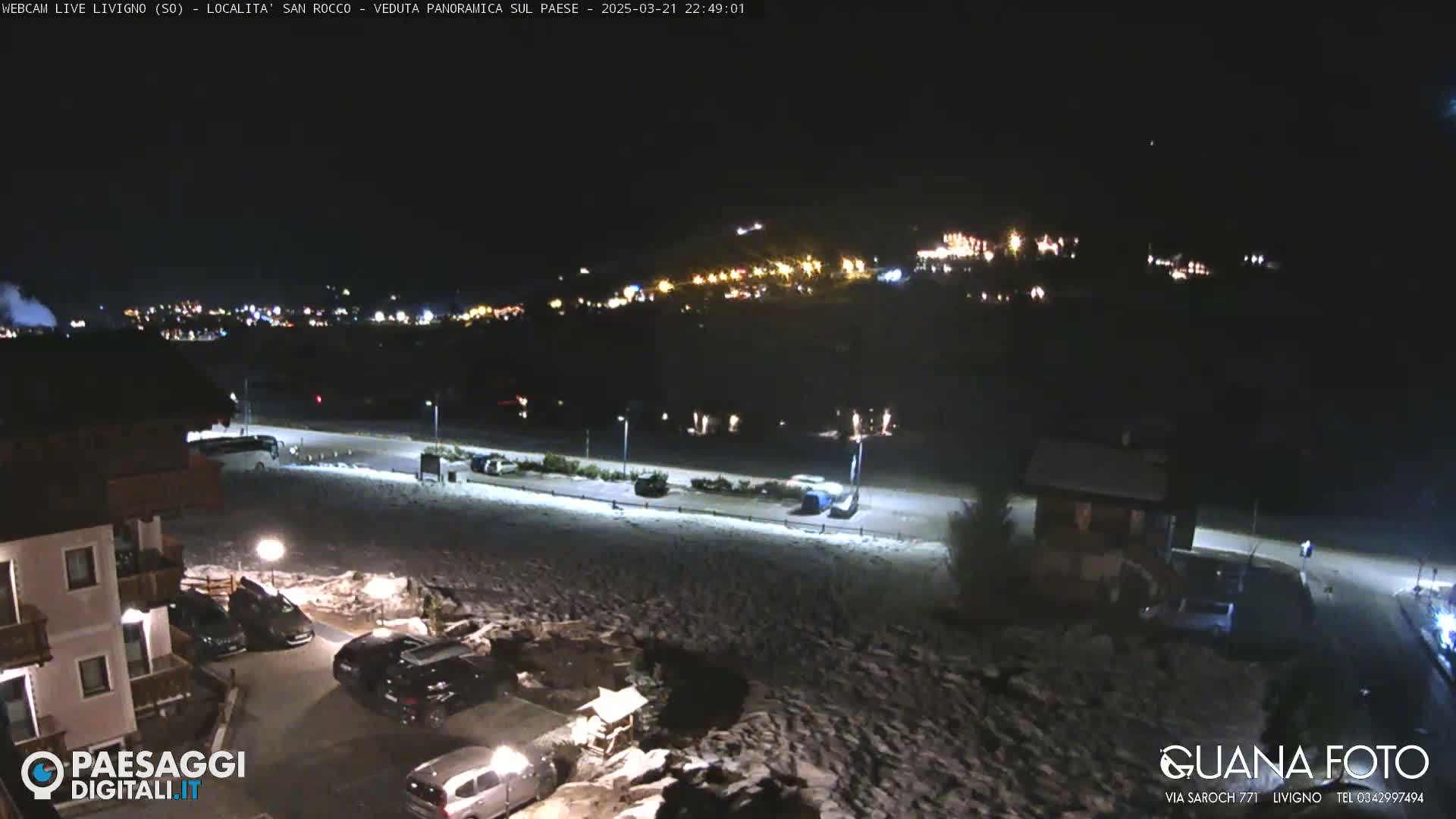 A nighttime, snowy scene shows a parking lot with several cars, a building, and a distant town illuminated with lights.