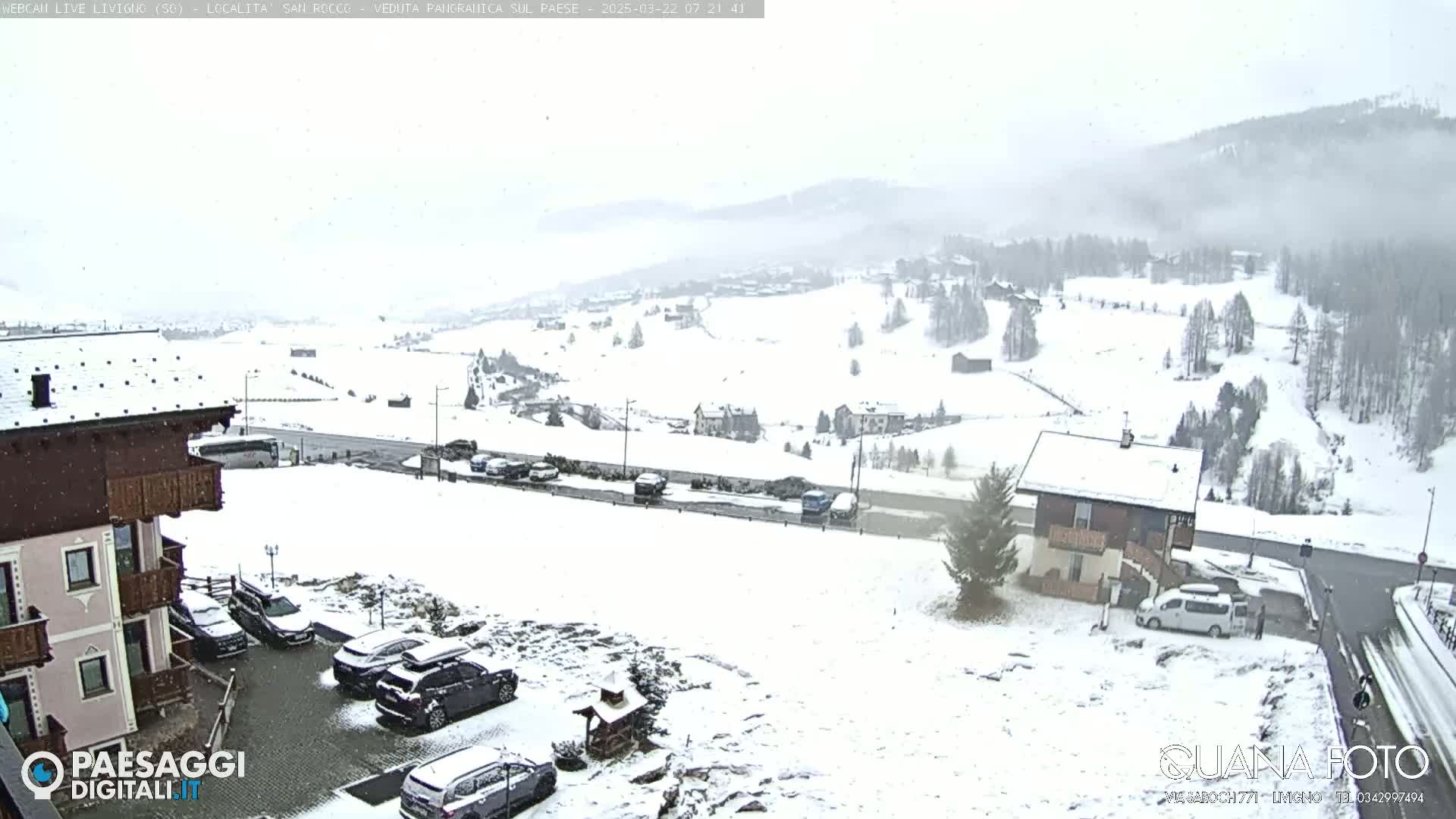 A snow-covered village in the mountains is lightly snowing, with cars parked in a lot near a building.