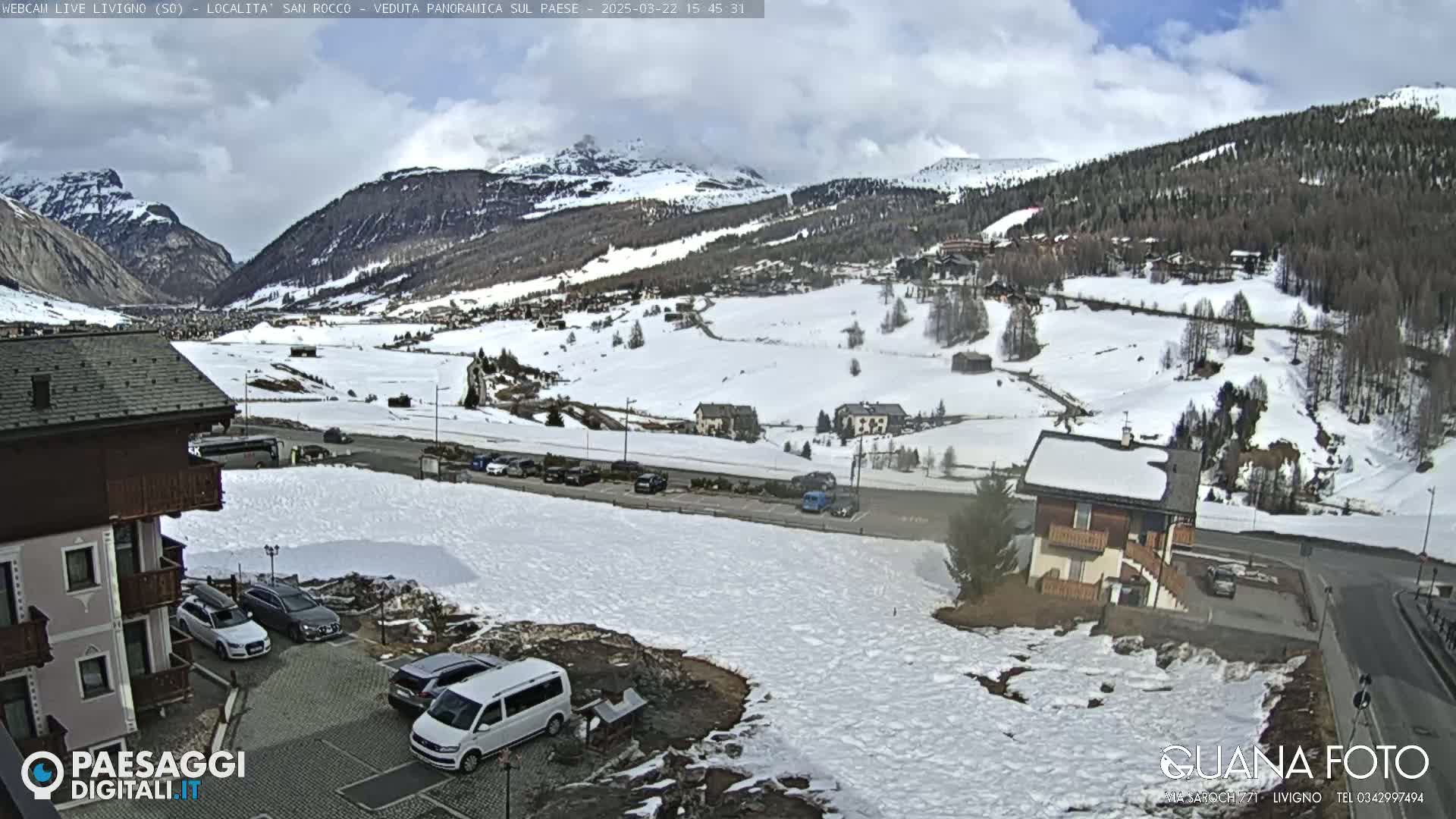 A partly cloudy day reveals a snow-covered mountain valley town with cars parked near buildings and a road running through it.