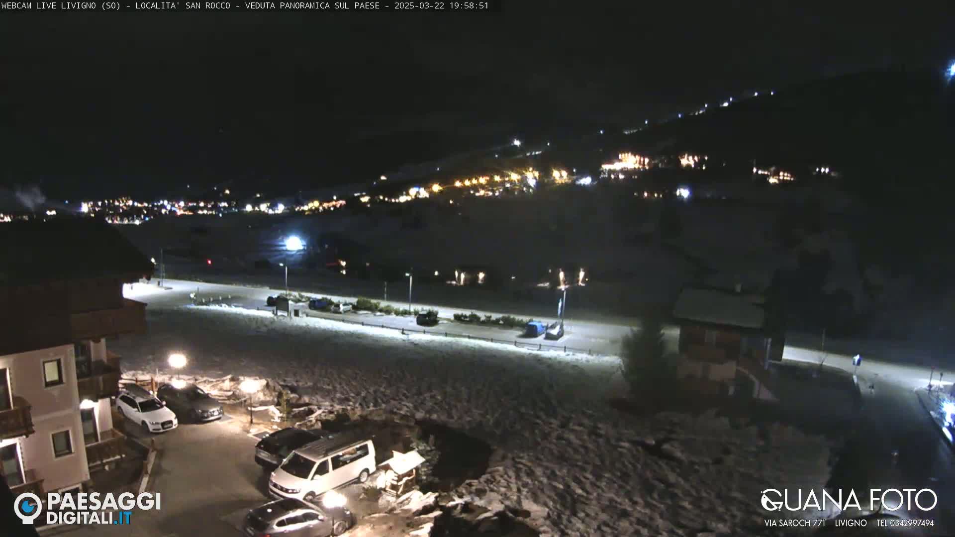A nighttime, snow-covered scene shows a village with illuminated buildings nestled in a hillside, viewed from an elevated position with cars parked nearby.