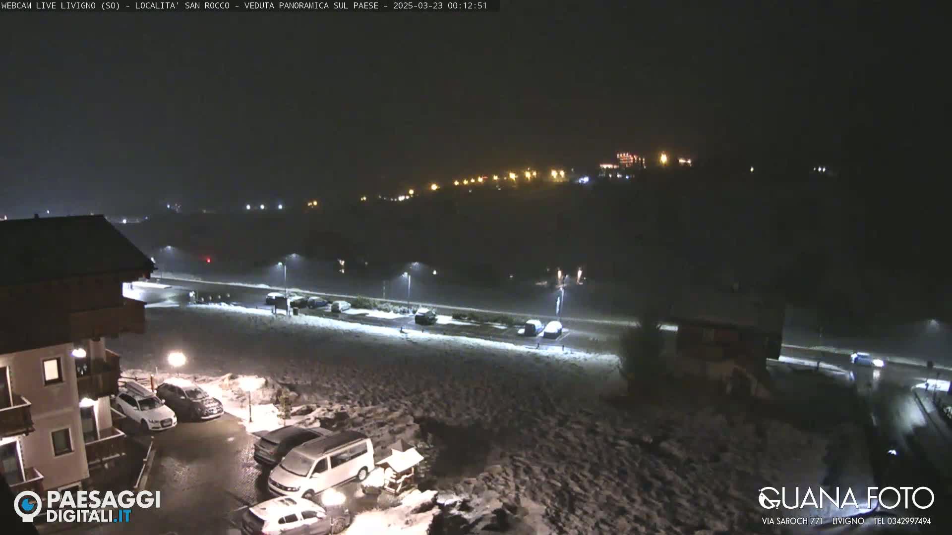 A nighttime, snow-covered parking lot and road are illuminated by streetlights, with buildings and distant lights visible in the background.