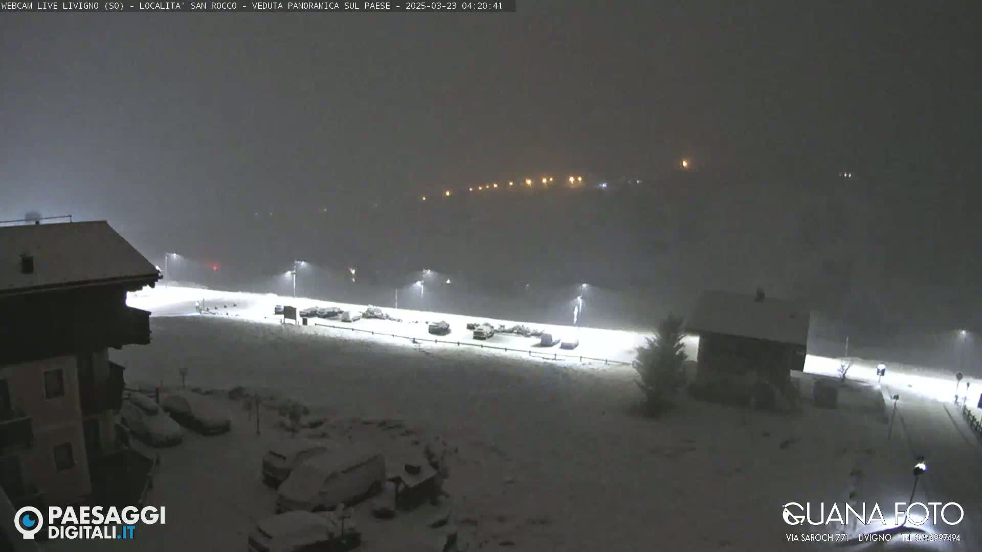 A snow-covered parking lot and buildings are illuminated by streetlights at night during a snowfall.