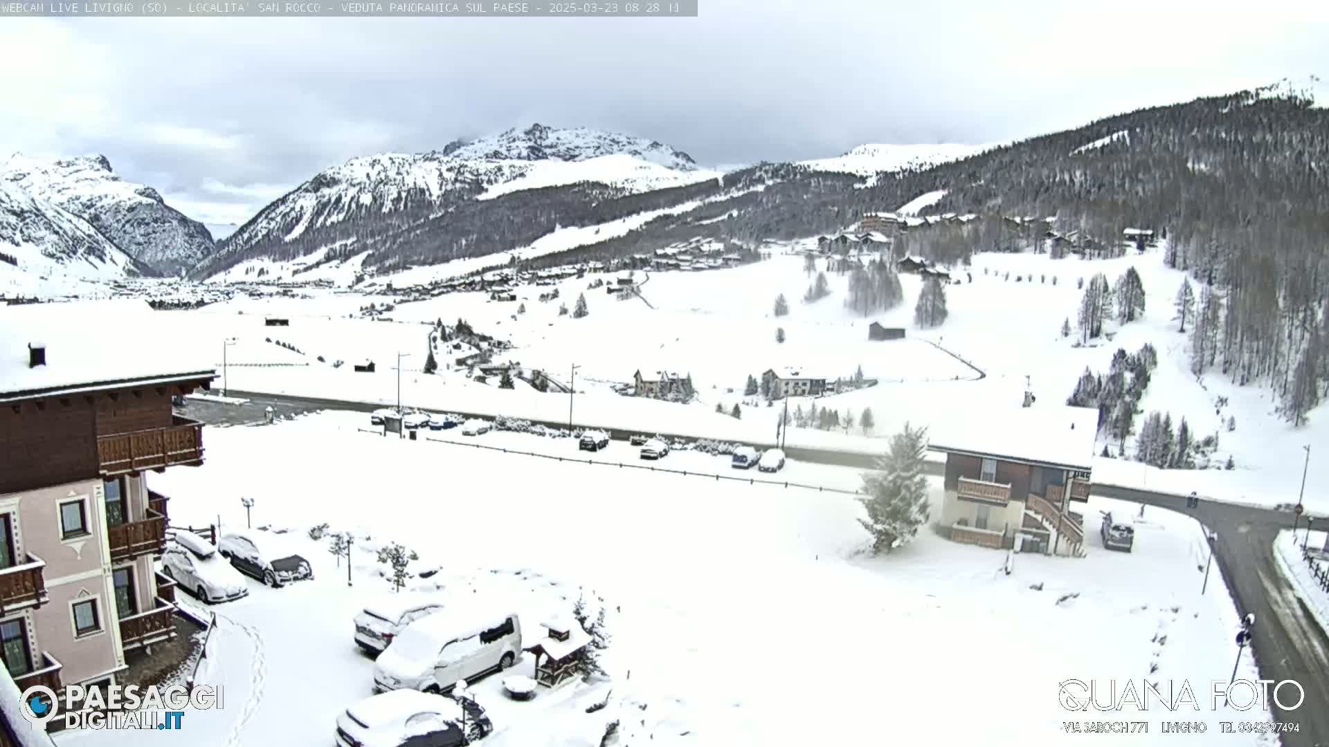 A snow-covered mountain village scene with overcast skies, featuring buildings, parked cars, and roads.