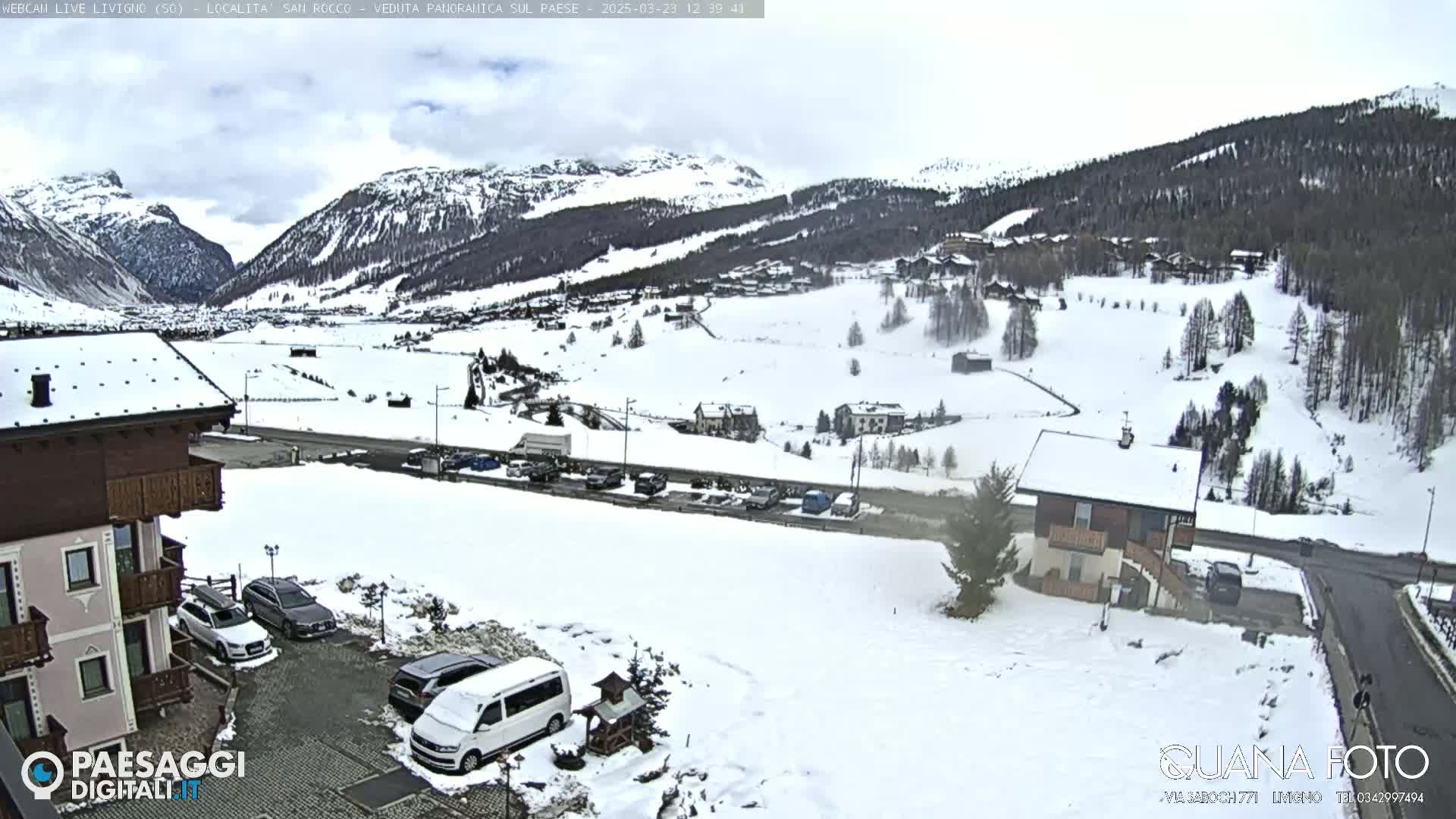 A snow-covered mountain valley village scene is visible from a slightly elevated vantage point, with cars parked in a lot near a building and overcast skies.